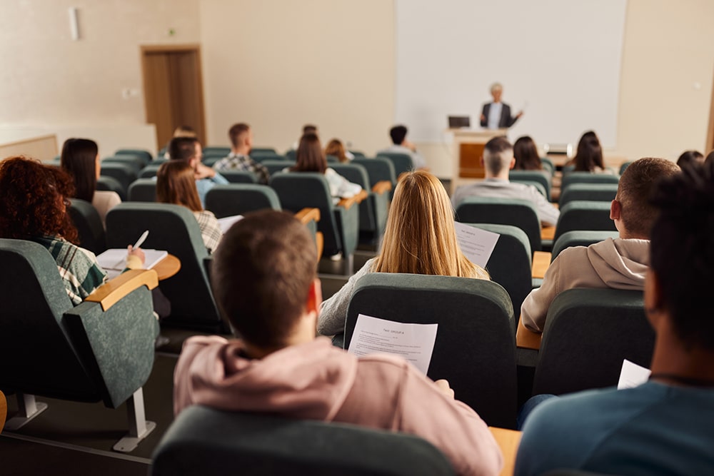 En föreläsningssal full med studenter sedda bakifrån. Längst fram står en lärare i ofokus.