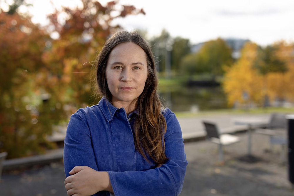 Porträtt på Jennie Brandén i halvprofil, ståendes framför ett grönområde med höstfärgade träd i bakgrunden.