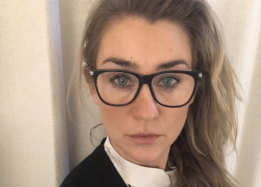 Porträttbild på Hanna Harnesk Hjortsberg, huvud och axlar, ståendes framför ett ljusgrått draperi.