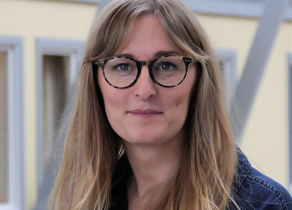 Porträttbild på Malin Joleby, huvud och axlar, ståendes framför en ljusgul husfasad.