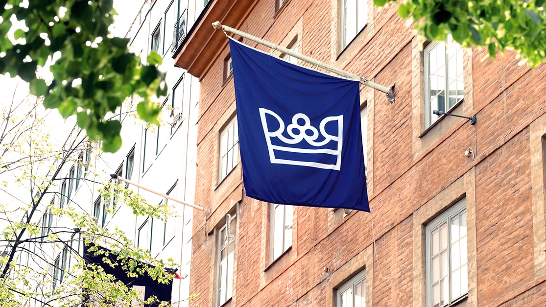 En stor blå flagga med Riksrevisionens vita logga hänger utmed en husfasad i ljusrött tegel.