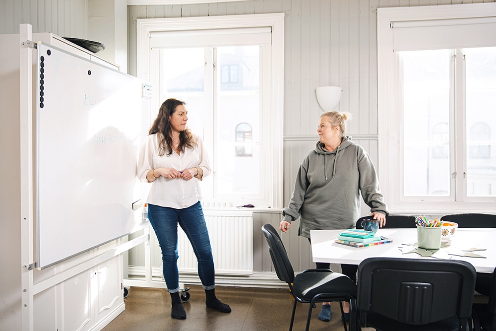 Två kvinnor stor i ett ljust mötesrum och pratar framför en whiteboard.