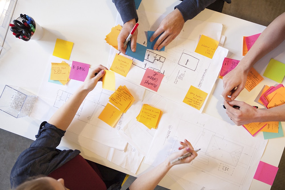 Tre personer samarbetar vid ett bord med ritningar och post-it-lappar, skriver och ordnar idéer med pennor.