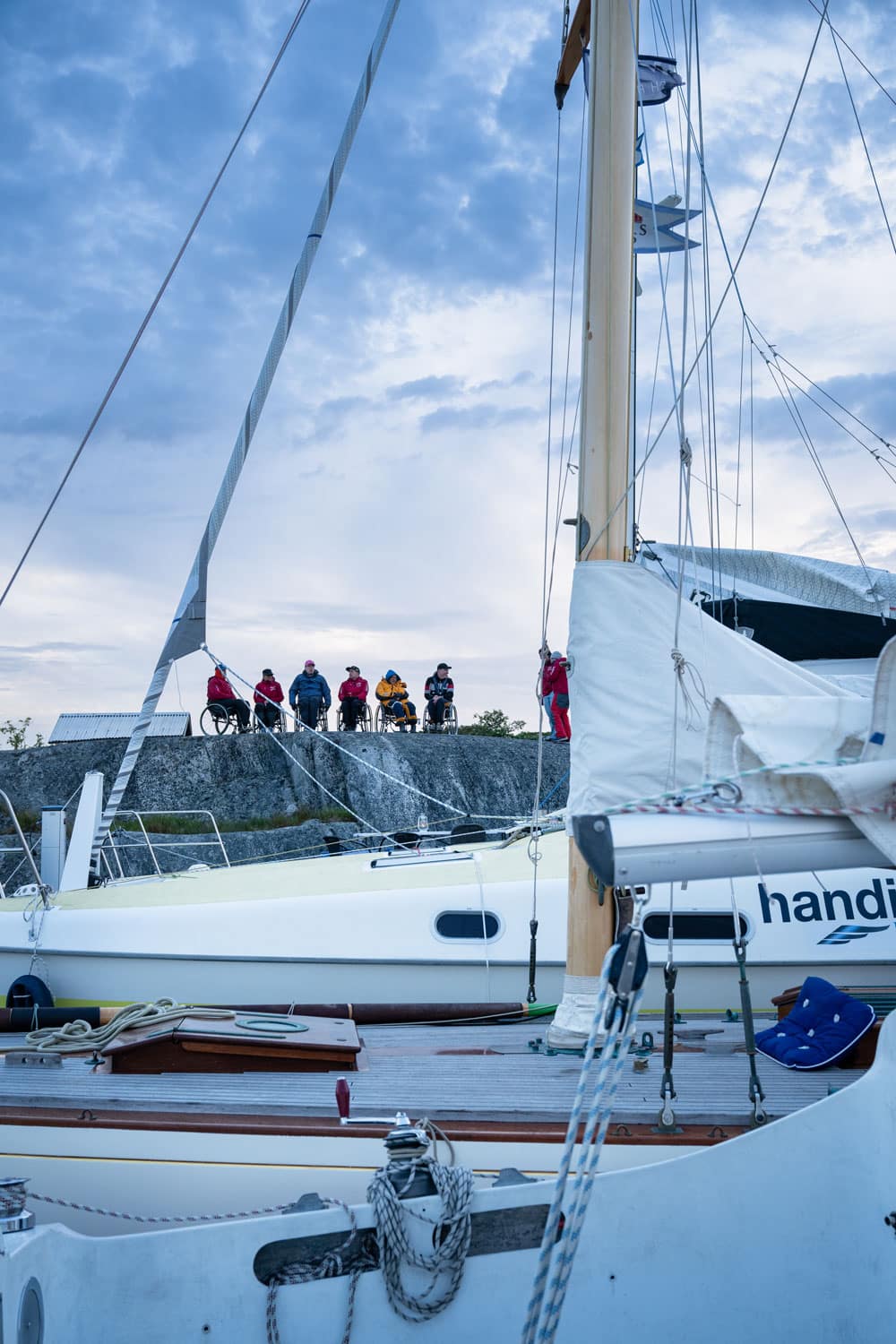 Sex personer i rullstol på en klippa med segelbåt i förgrunden.