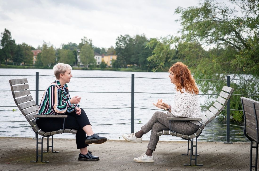 Jeanette Öhrlund och Erika Ottaeus sitter på träbänkar vid en sjö och pratar på en träaltan.