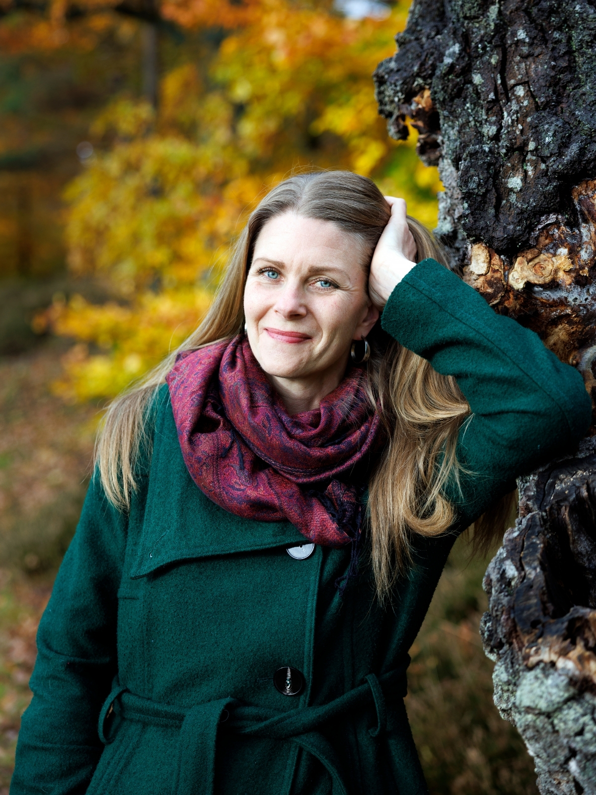 Marika Bjerstedt med långt hår och en röd halsduk lutar sig mot ett träd.