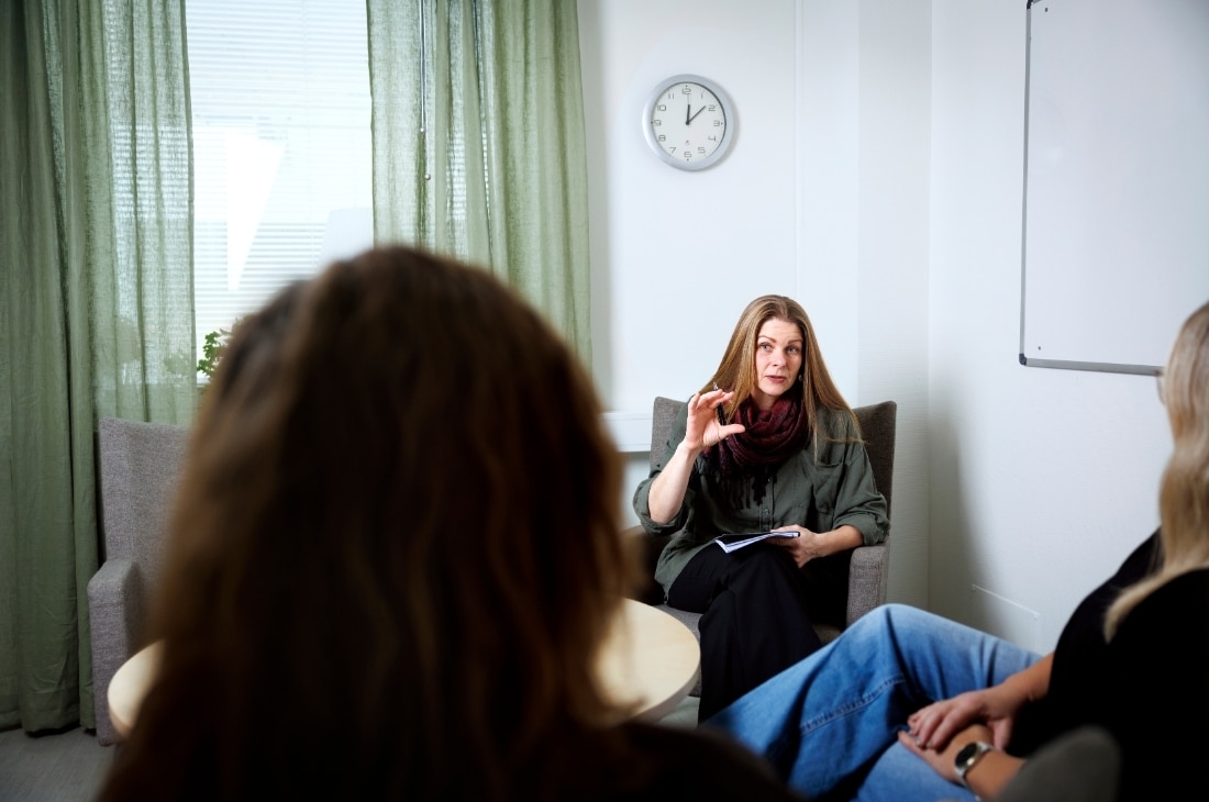 Marika Bjerstedt sitter på ett samtal; en kvinna talar och gestikulerar medan två lyssnar. En klocka och whiteboard syns bakom.