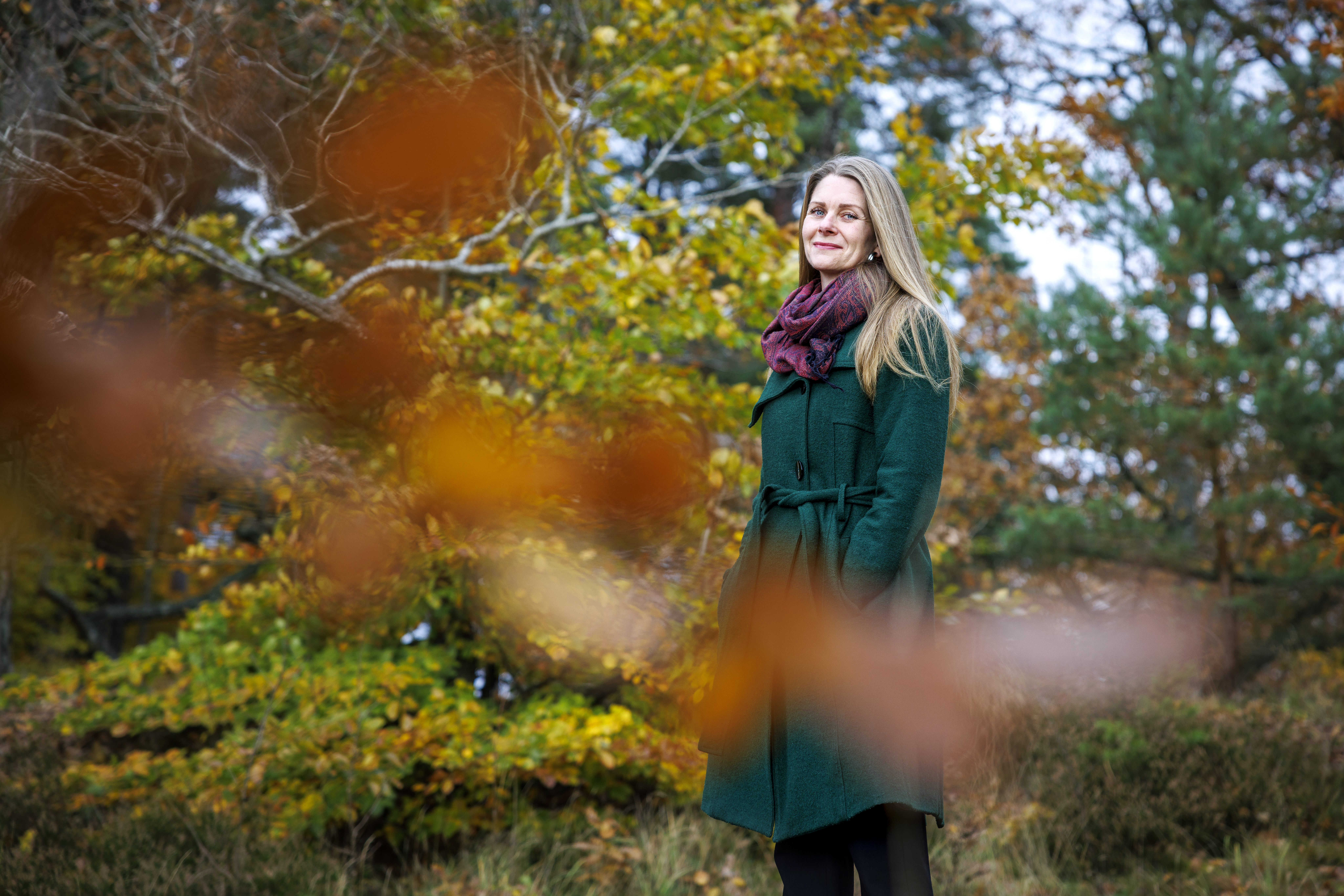 Marika Bjerstedt i grön kappa och vinröd halsduk står utomhus bland höstträd.