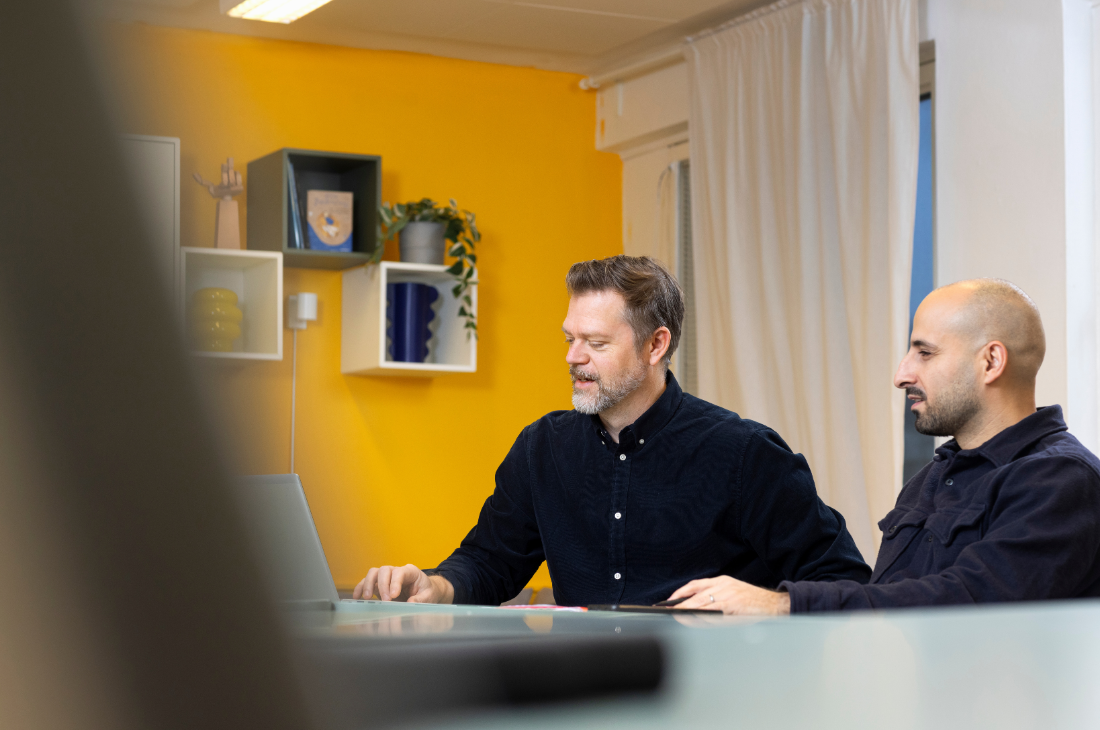 Andreas Wetterberg och kollega sitter vid ett bord på ett kontor och tittar på en datorskärm. I bakgrunden finns en gul vägg med hyllor och en växt.