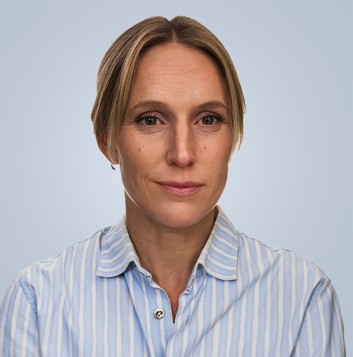 Porträttbild på Karin Persson med ljust hår iklädd en ljusblå och vitrandig skjorta. Tittar in i kameran och ler svagt.