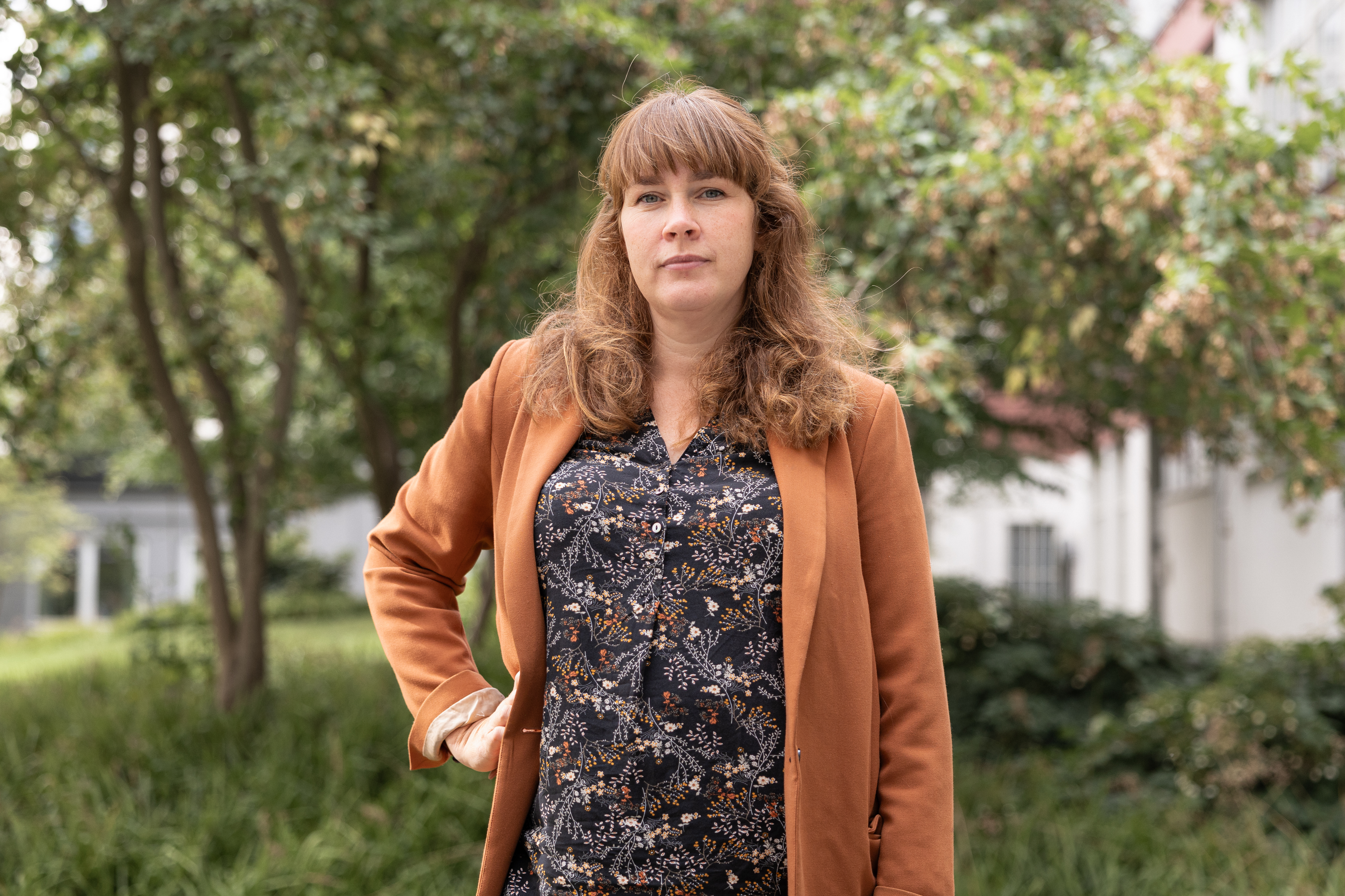 Johanna Azar med långt brunt hår står utomhus, har på sig en rostfärgad kavaj över en mönstrad blus, med växter och en byggnad bakom.