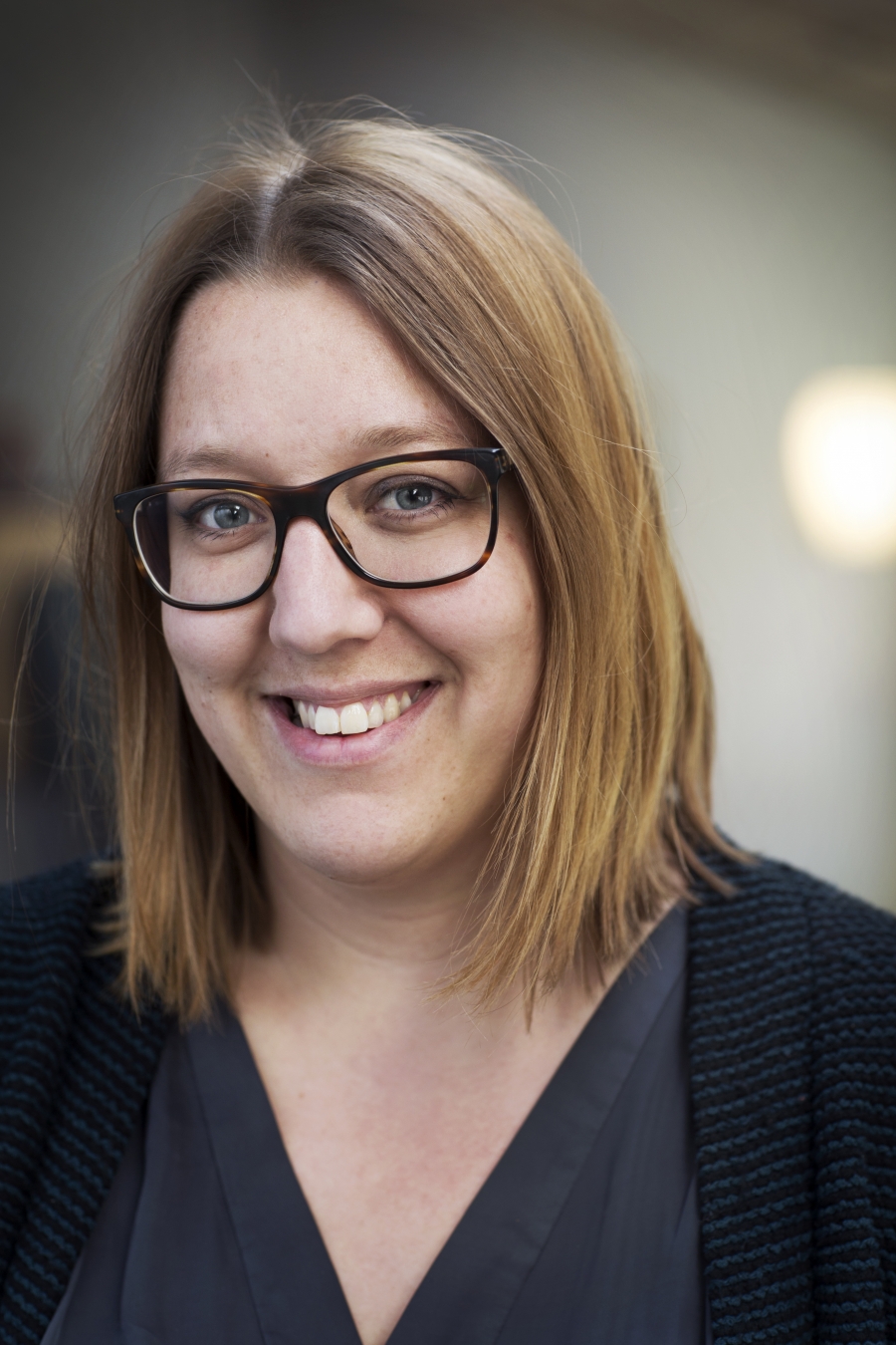 Anna Tegunimataka med axellångt ljusbrunt hår och glasögon, iklädd mörk topp och tröja, ser in i kameran mot en suddig bakgrund.