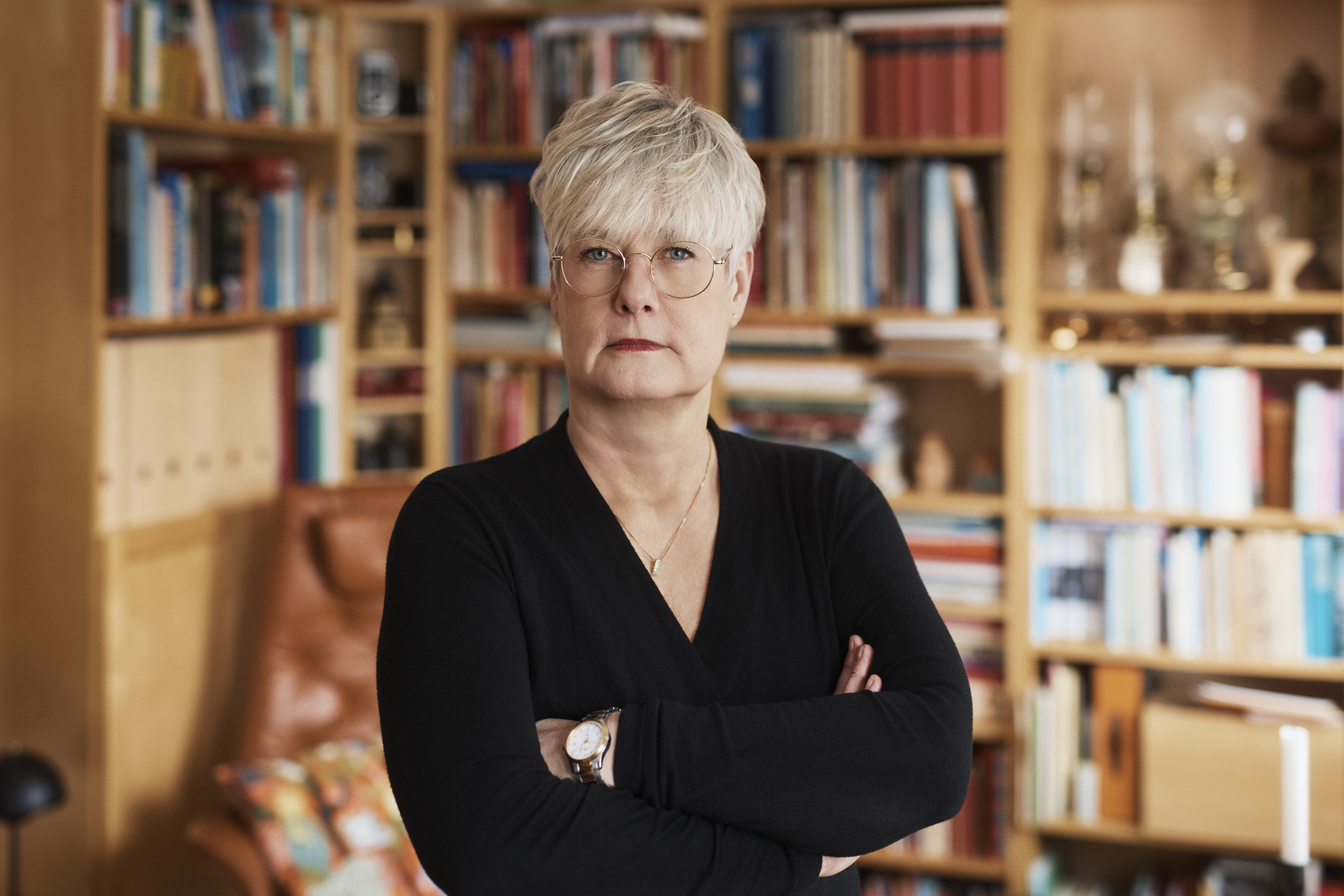 Marie Linder med kort ljusgrått hår och glasögon står med armarna i kors framför bokhyllor med böcker och prydnadssaker.