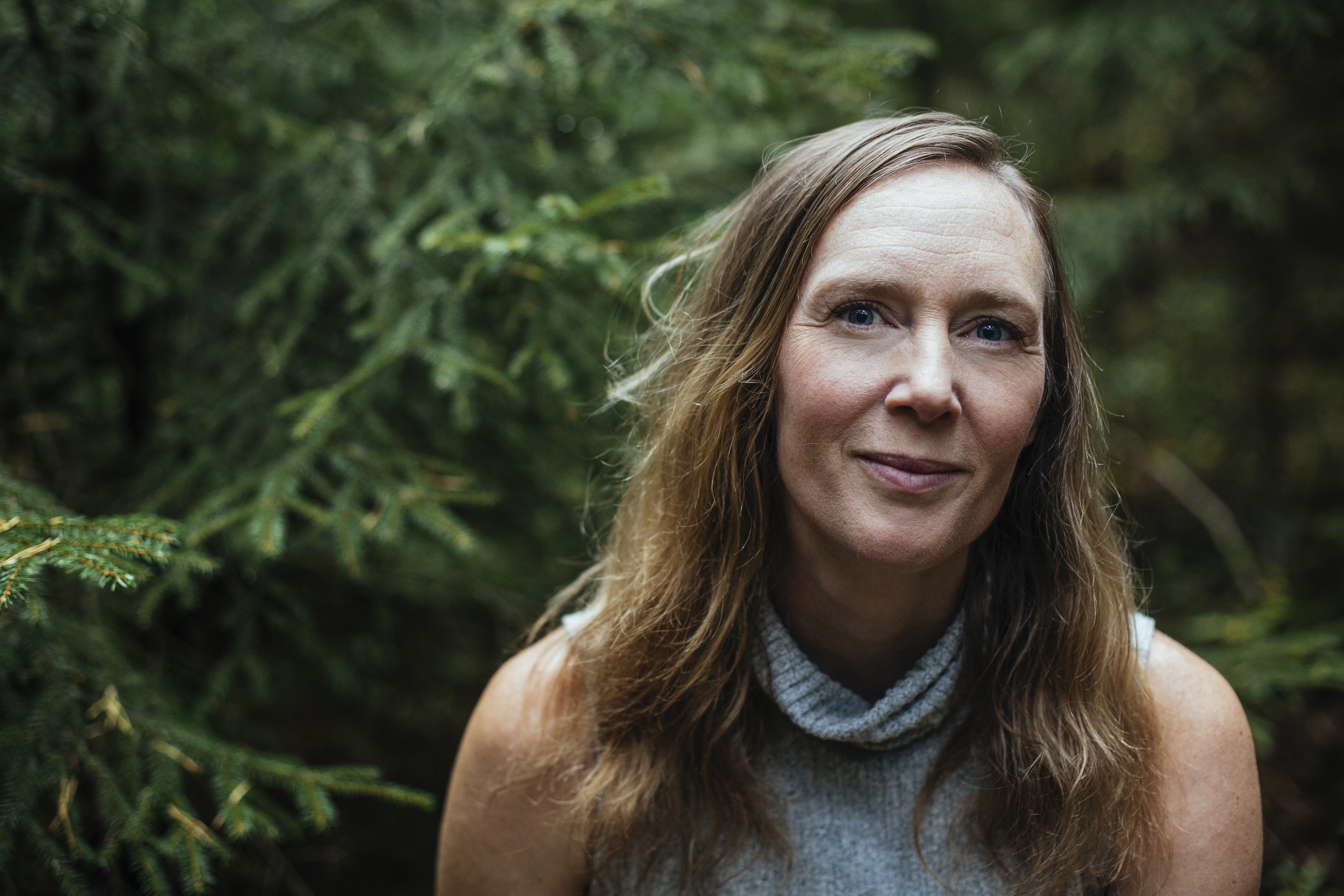 Jonna Bornemark med långt brunt hår och en grå, ärmlös topp står utomhus framför barrträd och tittar in i kameran.