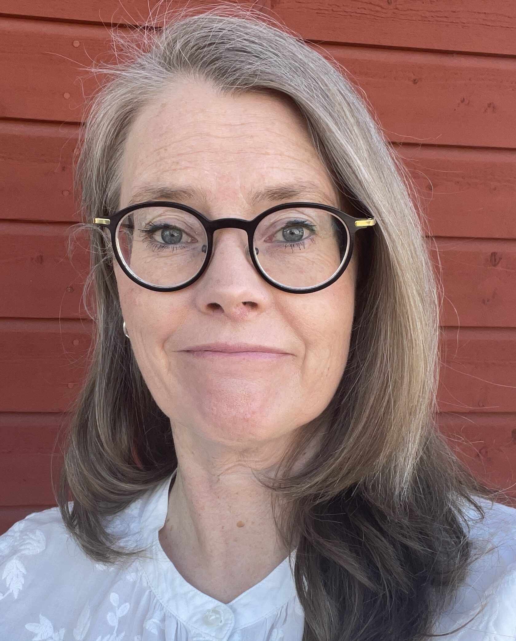 Lena Rehnholm med långt grått hår, runda svarta glasögon och en vit topp står framför en röd trävägg.