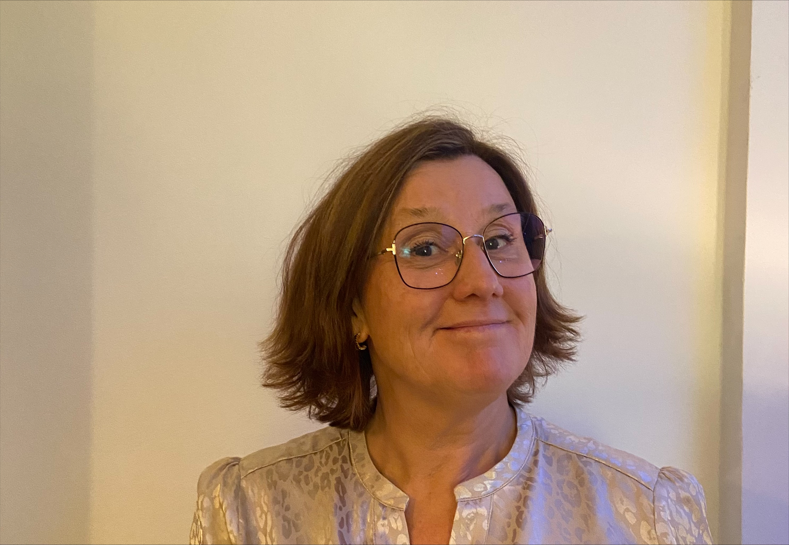 Cecilia Pihl med kort brunt hår och glasögon, klädd i en ljusmönstrad blus, står framför en ljus vägg och ler svagt.
