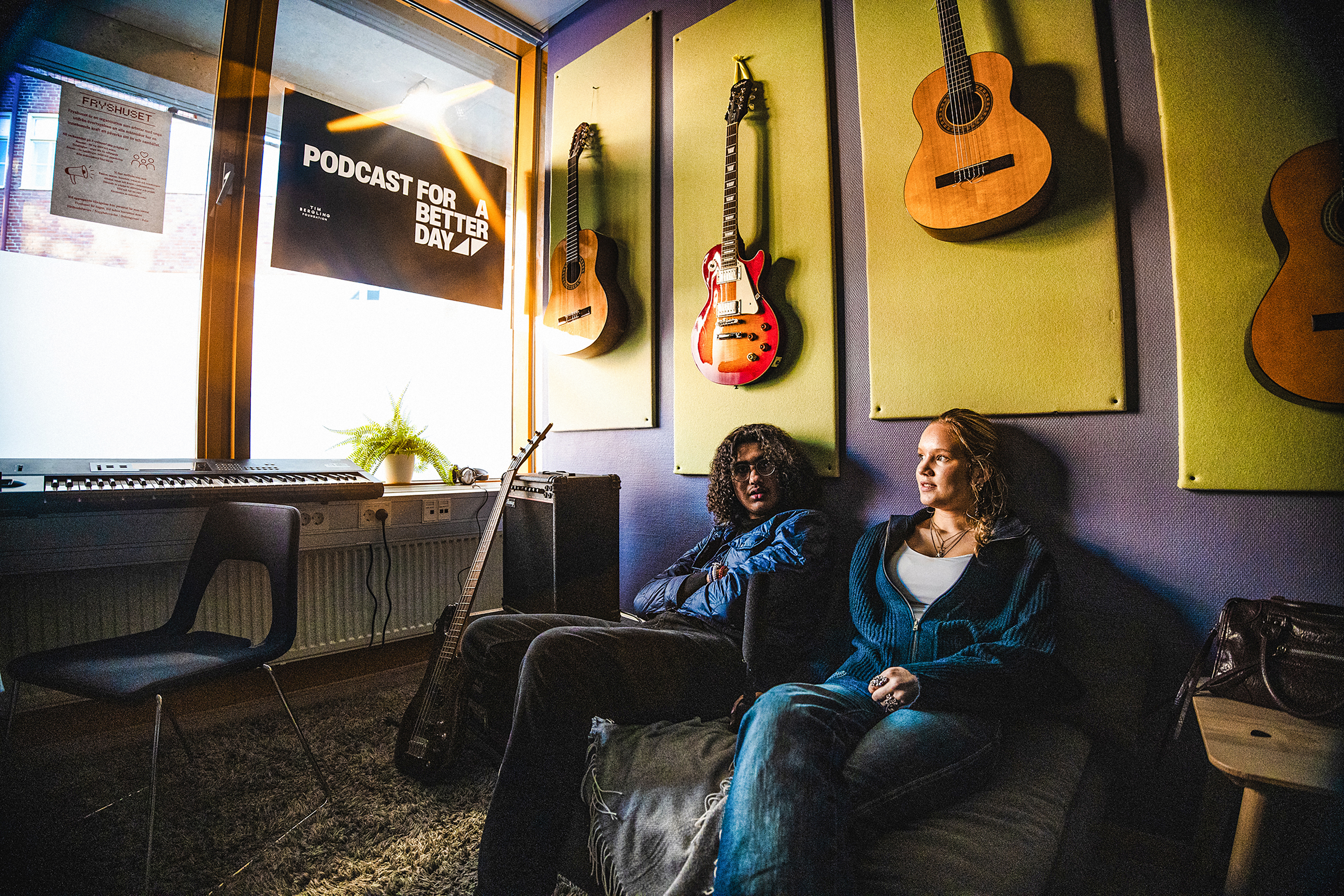 Lucas Pihl och Frances Carlberg sitter i en soffa i ett musikrum med gitarrer på väggen, ett keyboard vid fönstret och en skylt med texten ”Podcast for a Better Day”.