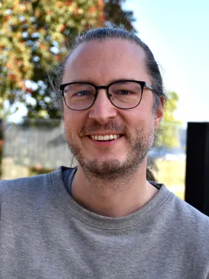 Erik Eriksson med glasögon och uppsatt långt hår bär en grå tröja och ler utomhus med träd och blå himmel i bakgrunden.