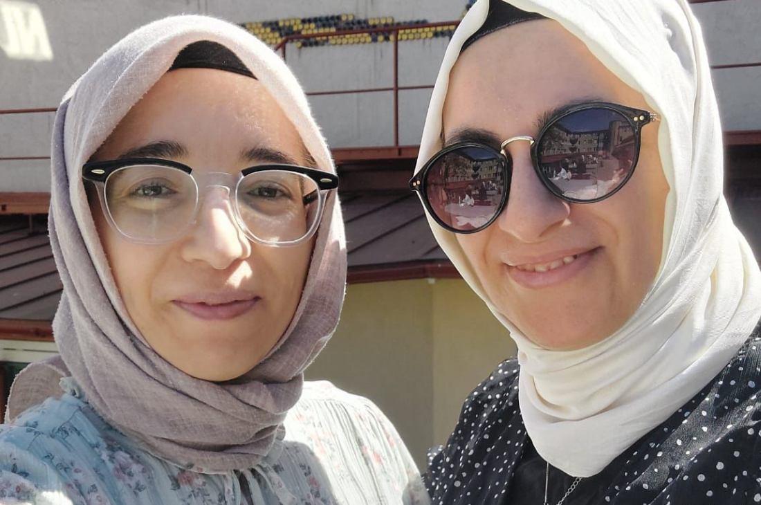 Två kvinnor med hijab och glasögon ler mot kameran utomhus.