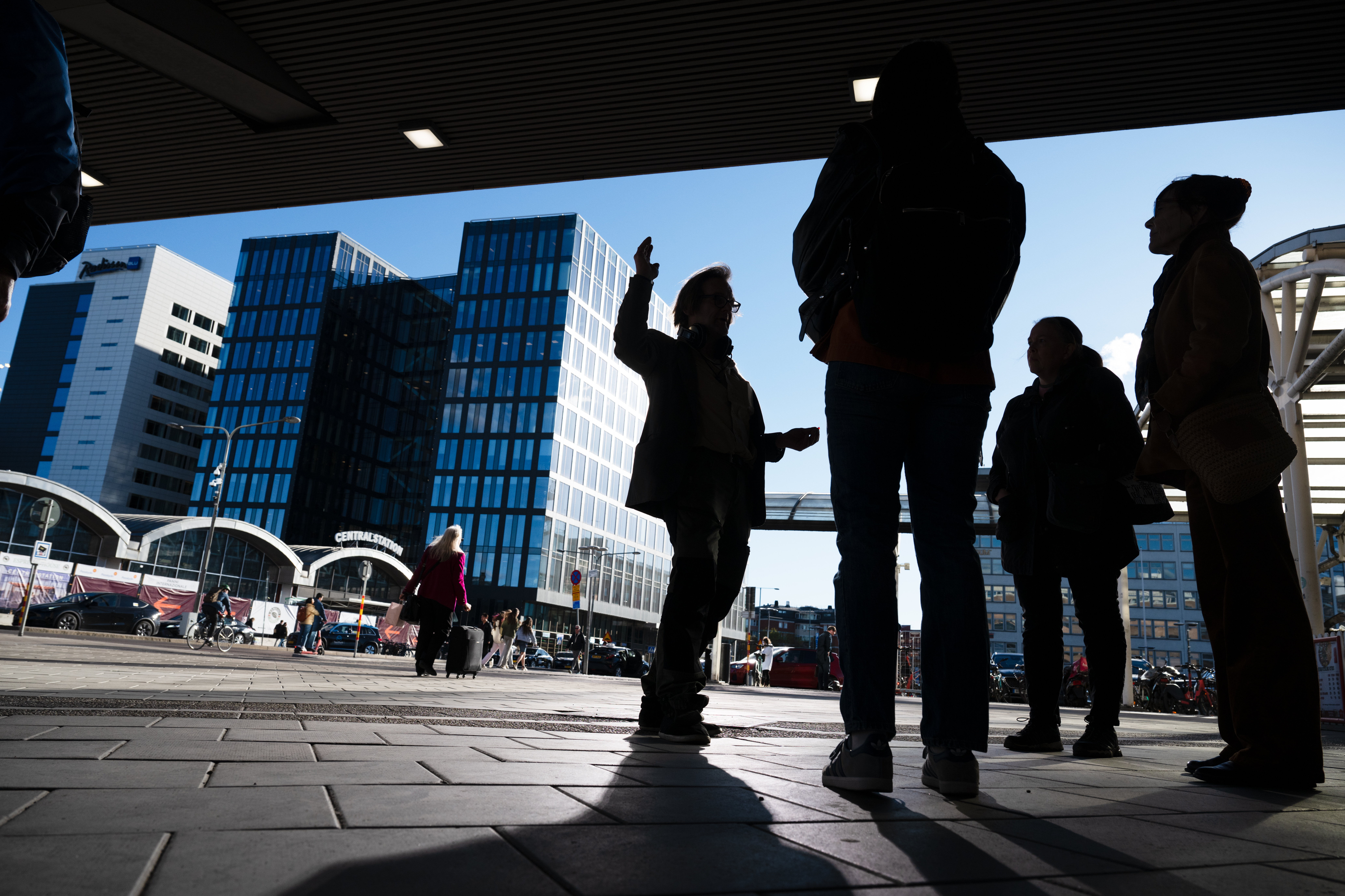En grupp människor står och pratar utanför moderna kontorsbyggnader, med skuggor på marken.