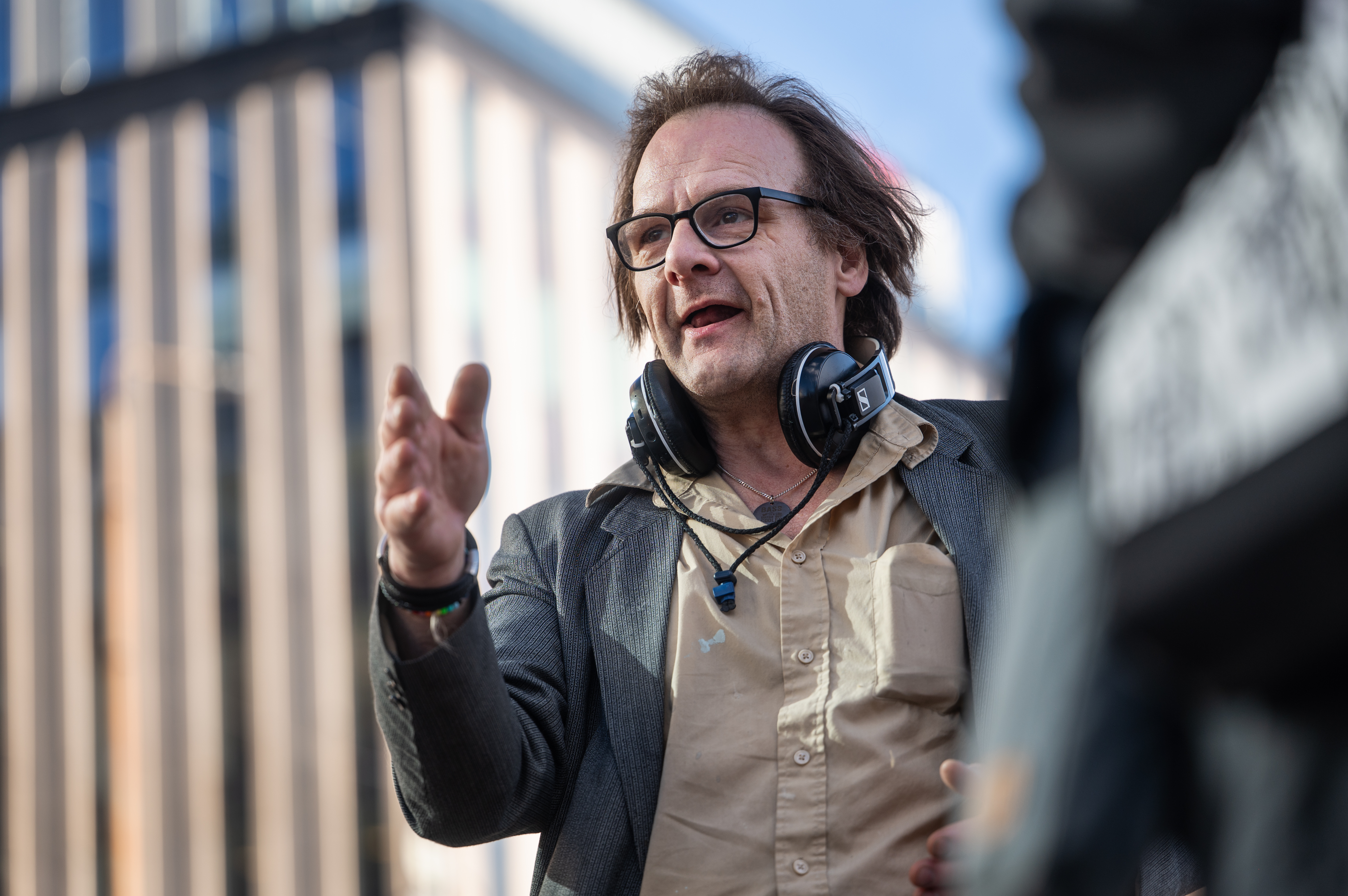 Hasse ”Hassebas” Karlberg med glasögon, hörlurar runt halsen och en beige skjorta gestikulerar med höger hand utomhus framför en modern byggnad.