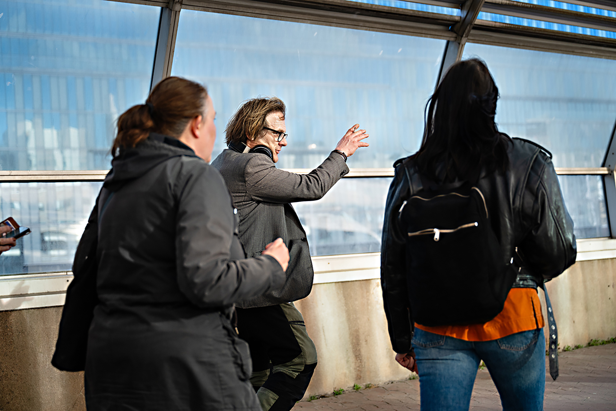 Tre personer befinner sig utomhus nära en glasvägg; Hasse ”Hassebas” Karlberg i mitten gestikulerar med handen medan hen pratar med de andra.