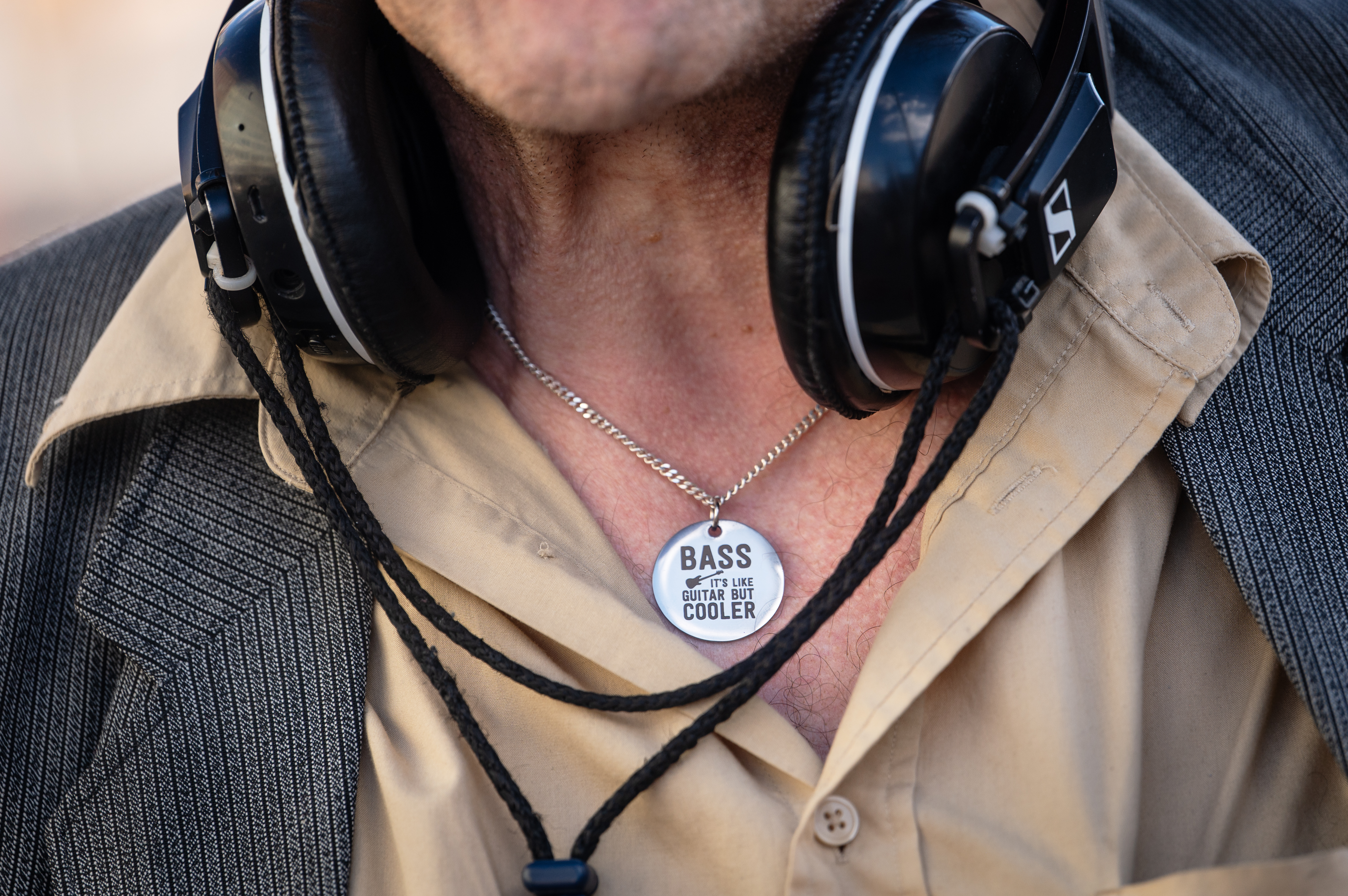 Hasse ”Hassebas” Karlberg klädd i en beige skjorta, kavaj och hörlurar runt halsen; ett halsband med en rund berlock där det står ”BASS OUT THE LIFE GUITAR JUST COOLER”.