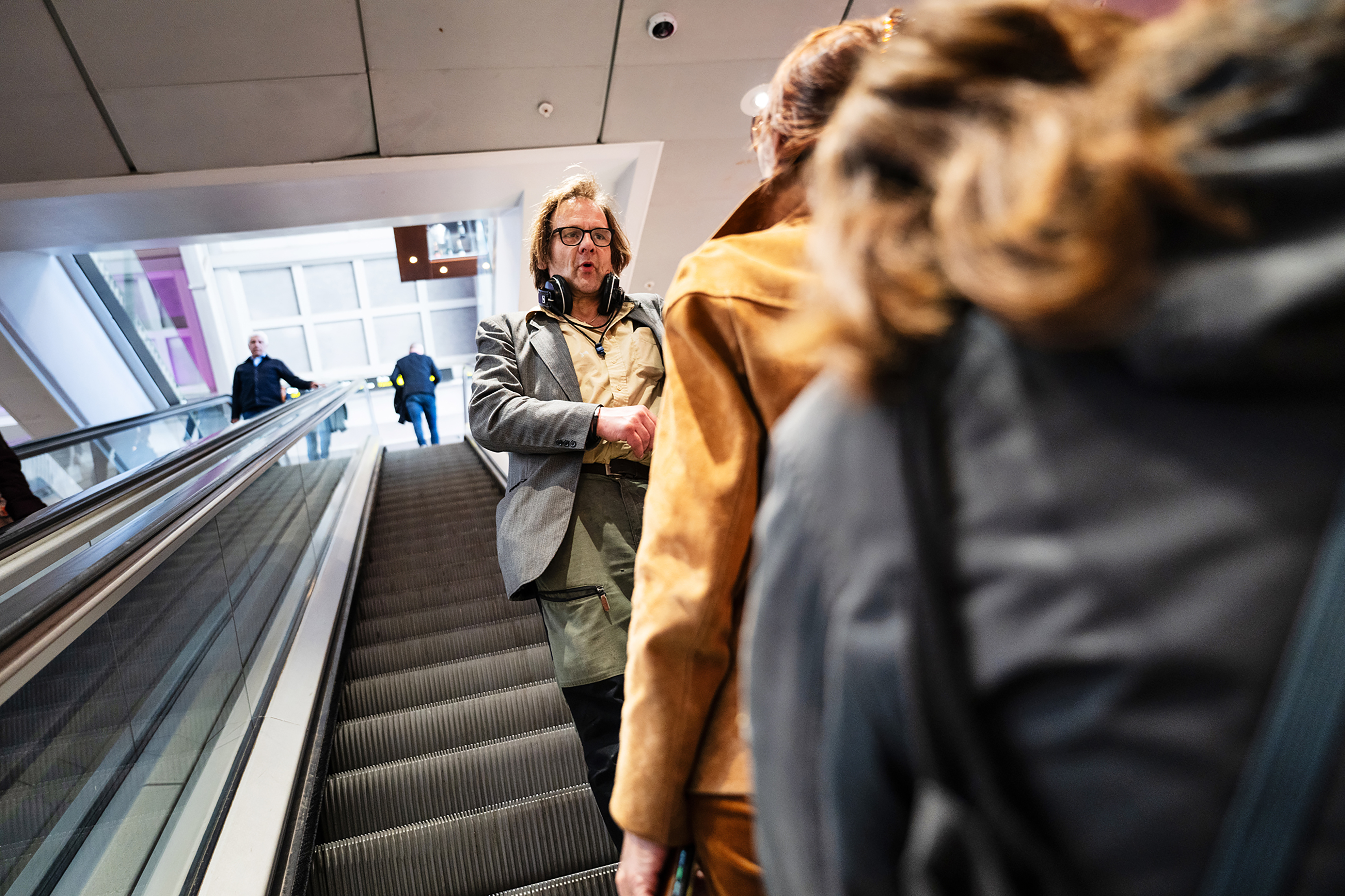 Hasse ”Hassebas” Karlberg med glasögon och hörlurar står på en rulltrappa som går uppåt, medan flera personer befinner sig på rulltrappan som går nedåt i en inomhusmiljö.