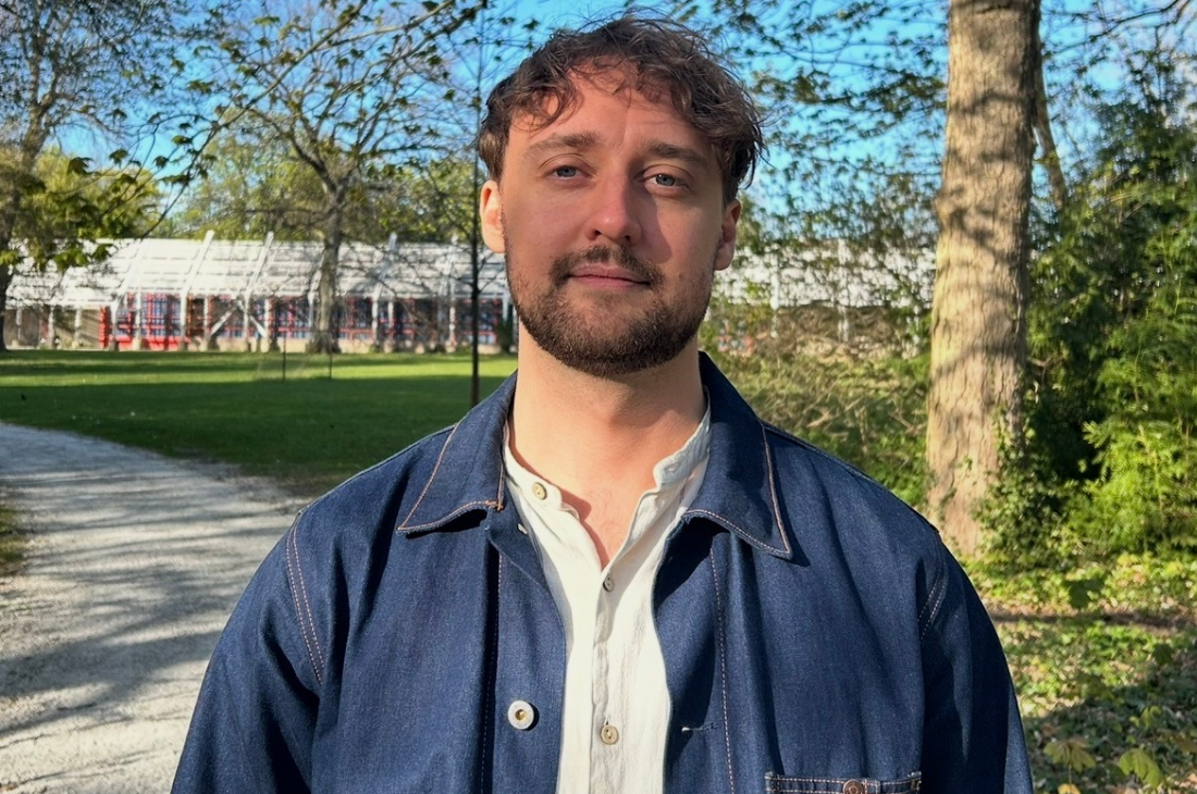 Jonathan Lundborg med kort brunt hår och skägg står på en gångväg i en park, klädd i en vit skjorta och en blå jacka. I bakgrunden syns träd och en glasbyggnad.