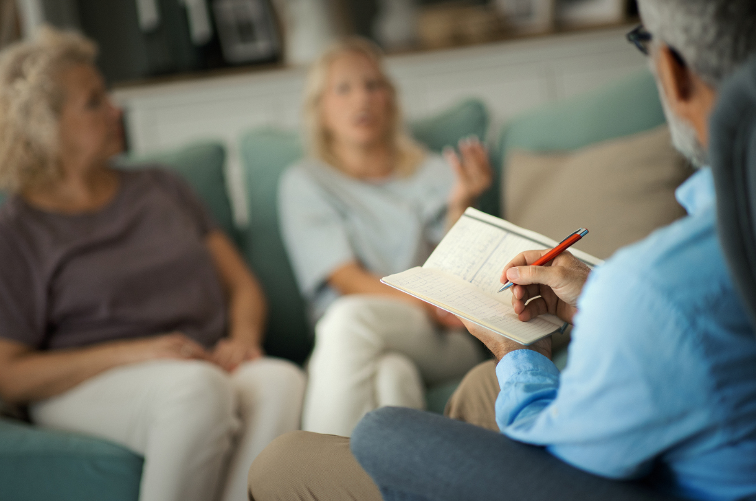 En terapeut gör anteckningar medan två kvinnor sitter i en soffa och pratar under en samtalssession i ett vardagsrum.