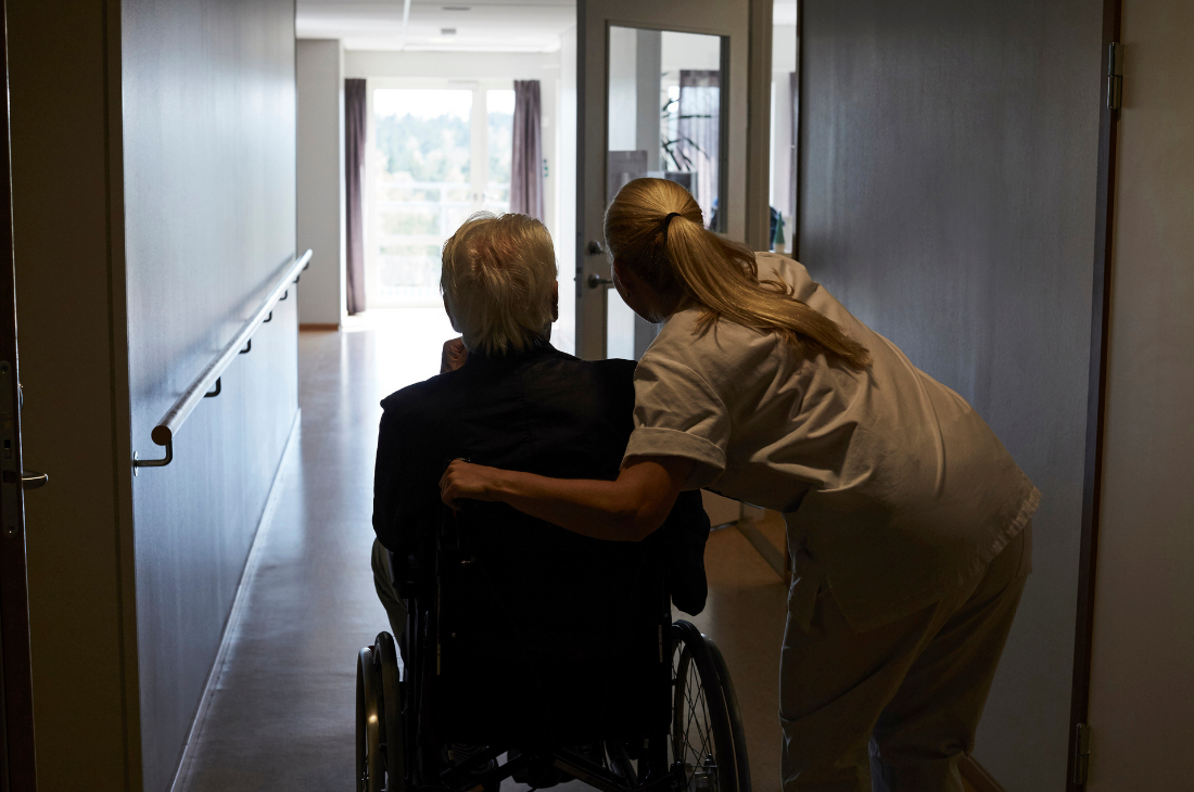 En vårdgivare står bredvid en äldre person i rullstol och tittar ut genom en öppen dörr i änden av en korridor på ett vårdhem.
