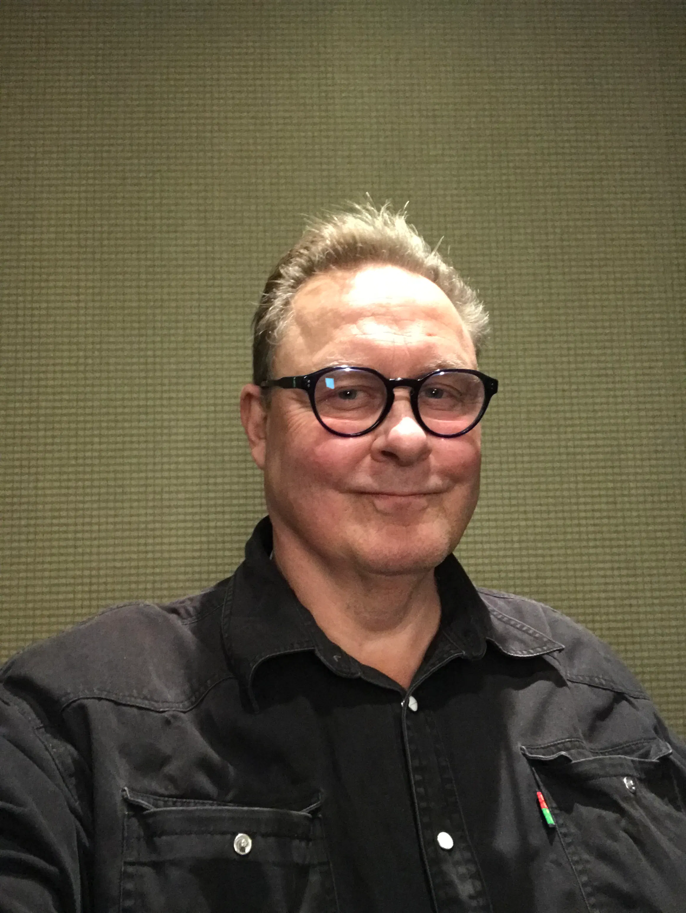 Kaj Gustafsson med glasögon och svart skjorta står framför en beige vägg, tittar in i kameran och ler svagt.