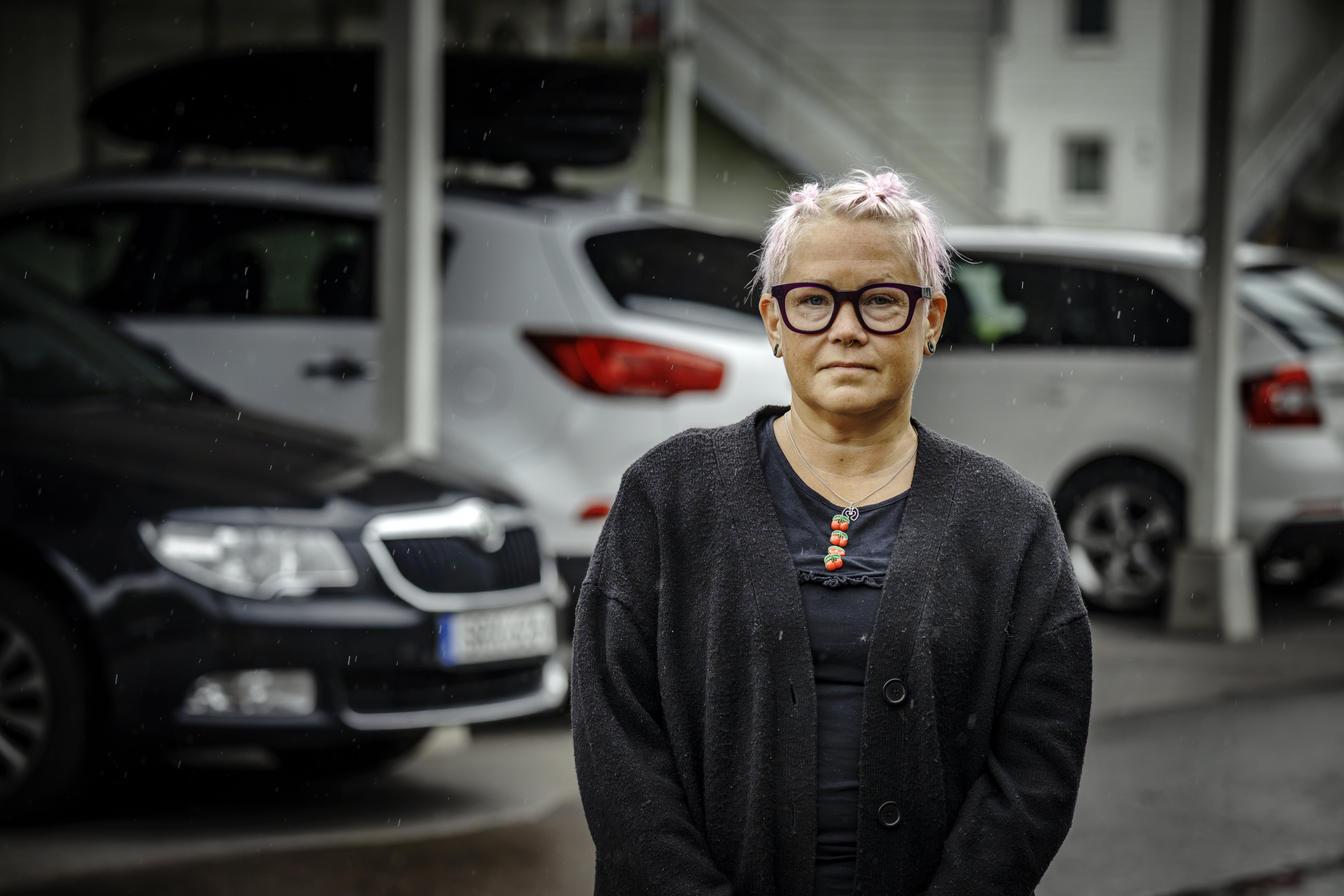 Marie Gustafsson med ljusrosa hår och glasögon står utomhus framför parkerade bilar en regnig dag. Hon har en svart tröja på sig och visar inga särskilda känslor.