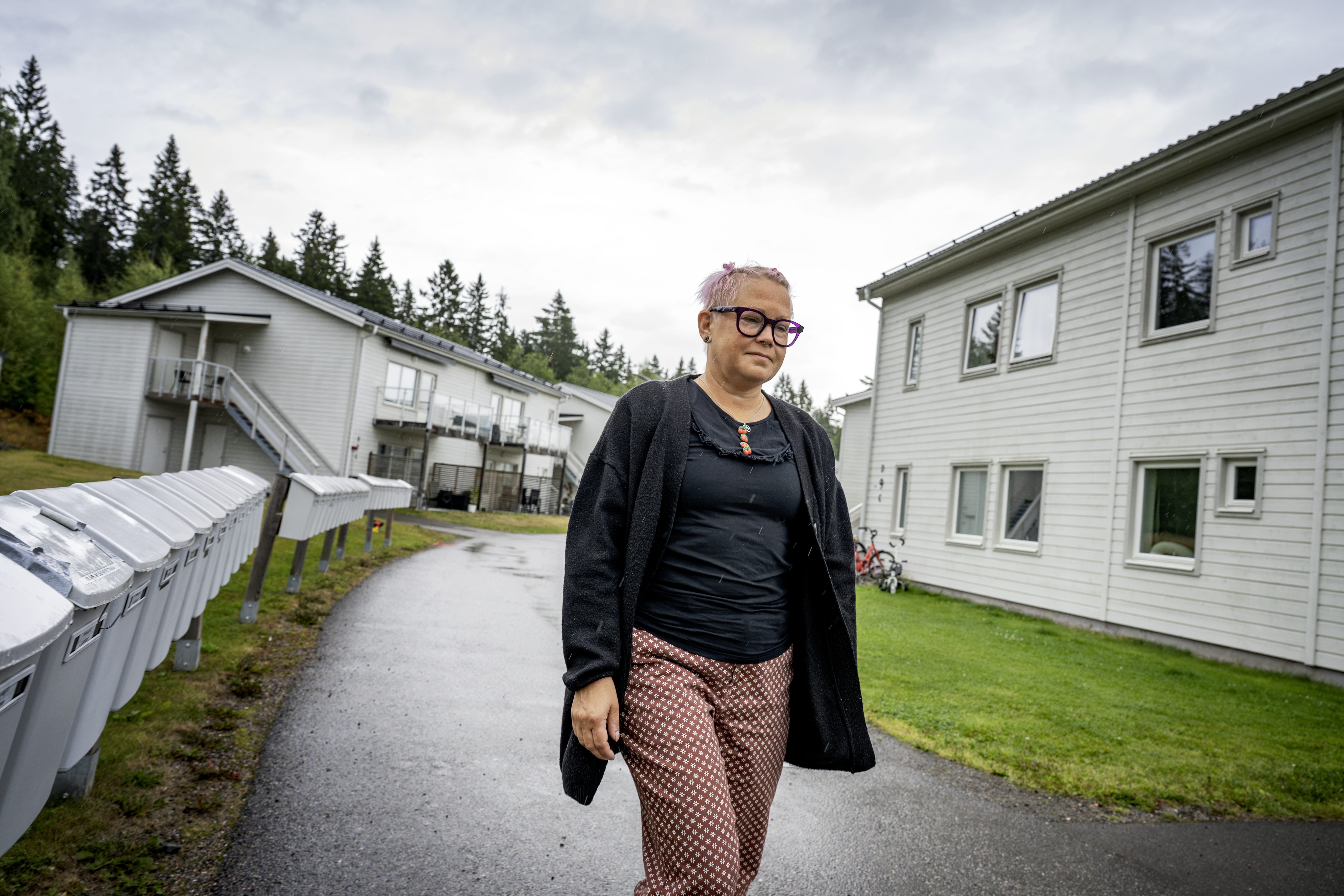 Marie Gustafsson med rosa hår och glasögon går på en asfalterad gångväg mellan ljusa bostadshus en molnig dag. I bakgrunden syns gräs och träd.