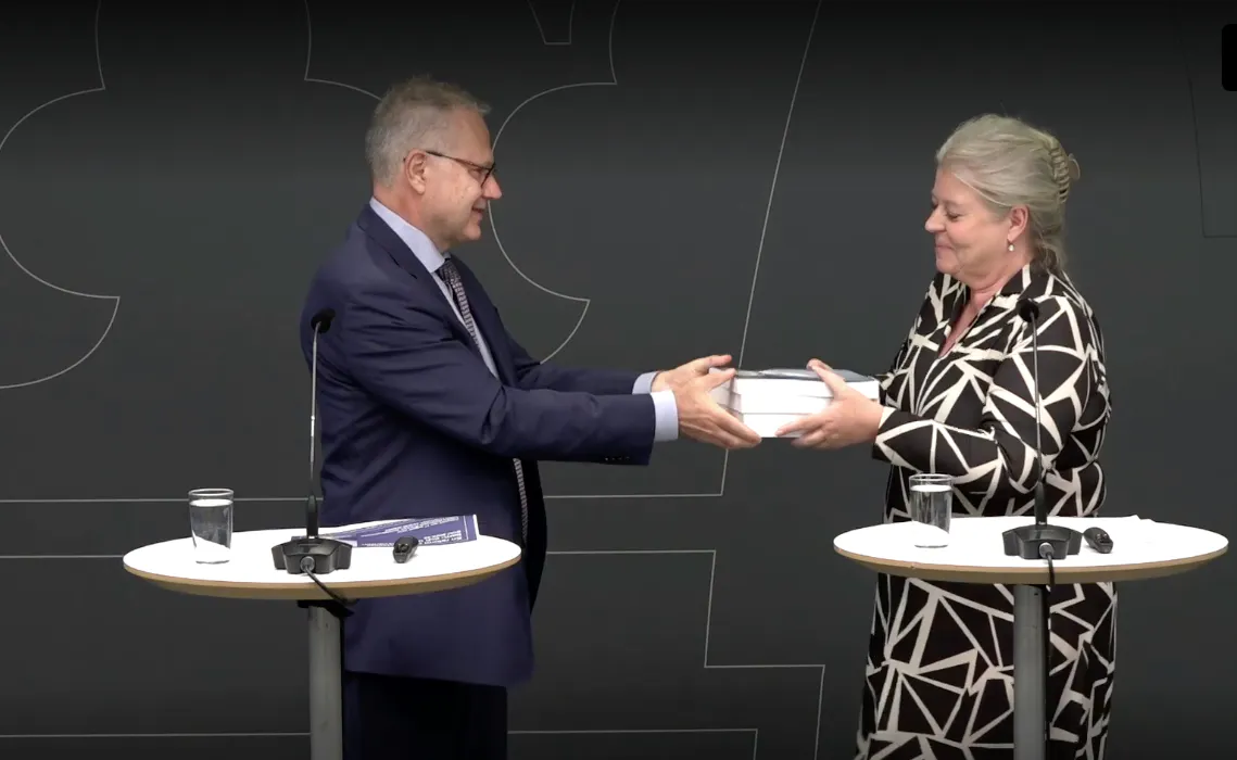 Stefan Holgersson i kostym överlämnar en bunt dokument till socialtjänstminister Camilla Waltersson Grönvall i klänning vid ett bord med mikrofoner och vattenglas.