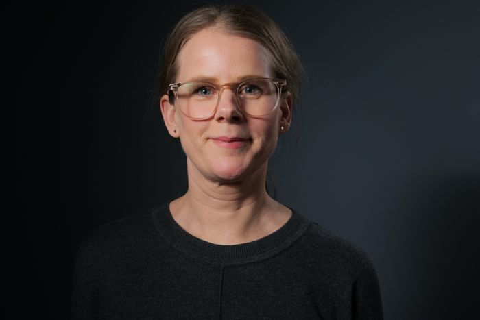 Eva-Lotta Nilsson med glasögon och uppsatt ljusbrunt hår, klädd i en mörk tröja, står framför en mörkgrå bakgrund och tittar in i kameran med ett neutralt ansiktsuttryck.