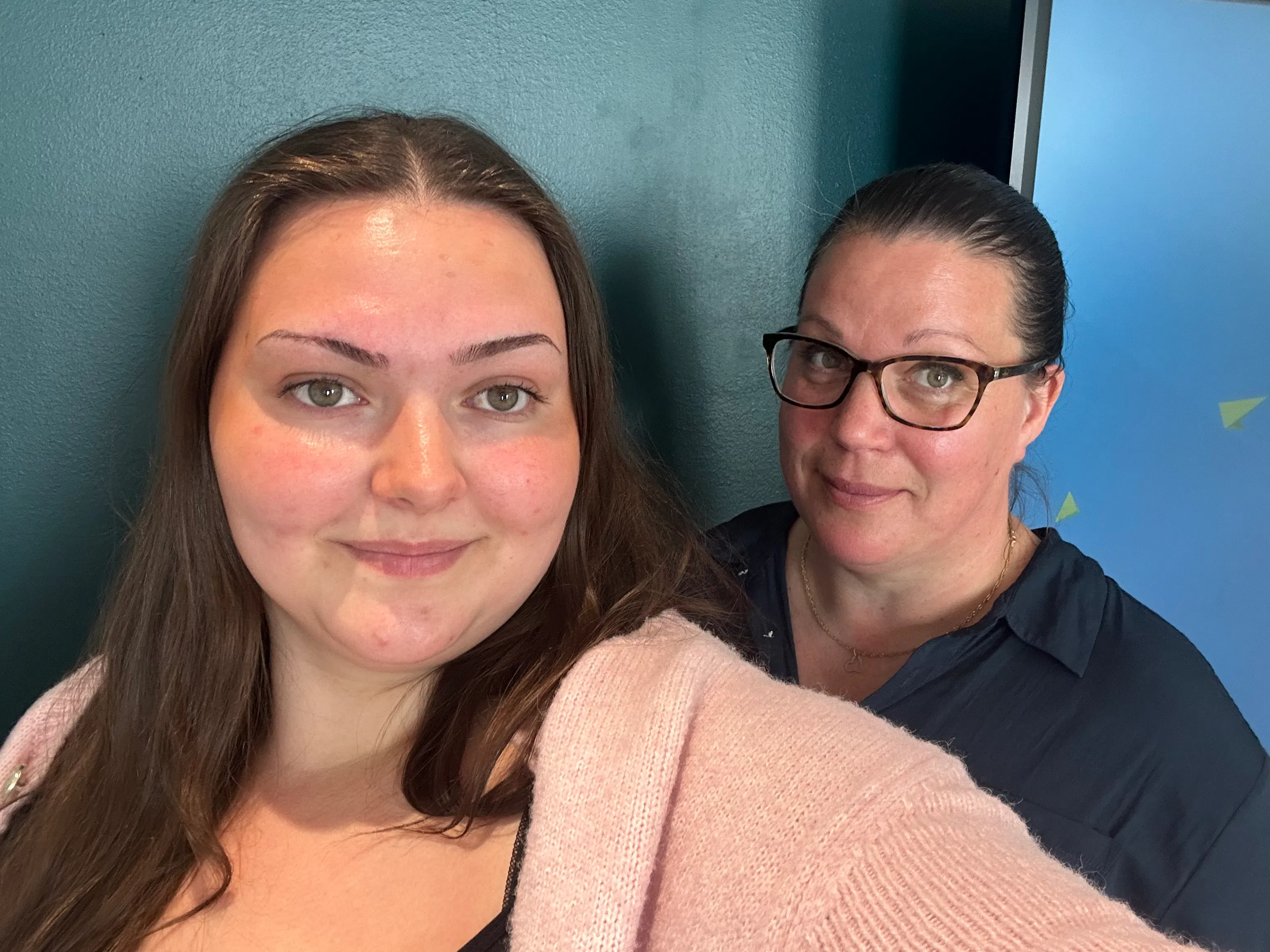 Victoria Fulici och Åsa Christiernin står inomhus, en längst fram i en rosa tröja och en bakom med glasögon och en mörk tröja; båda vänder sig mot kameran framför en turkos vägg.
