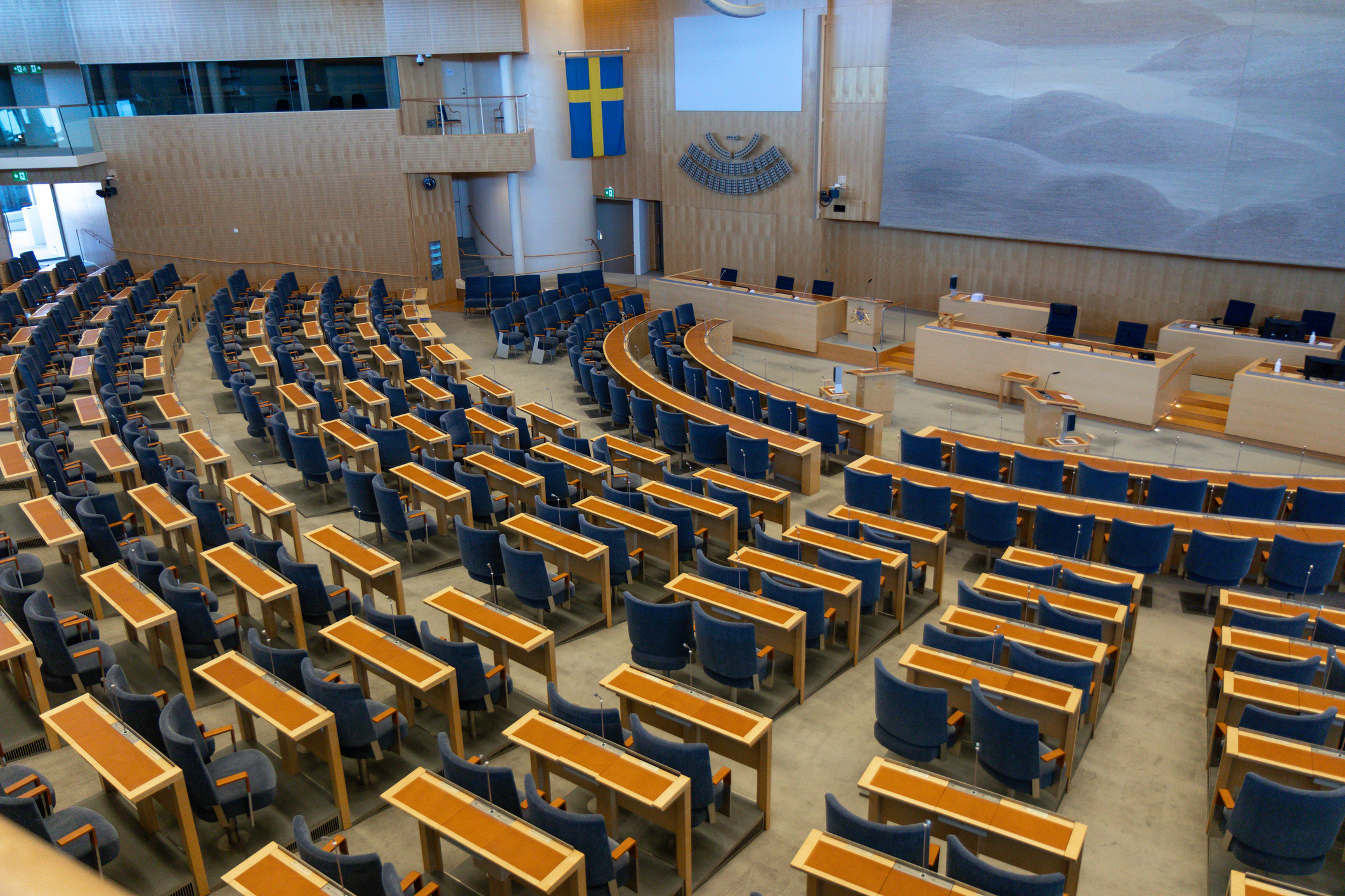 En tom parlamentssal med rader av träbord och blå stolar, ett talarstativ och en svensk flagga längst fram.