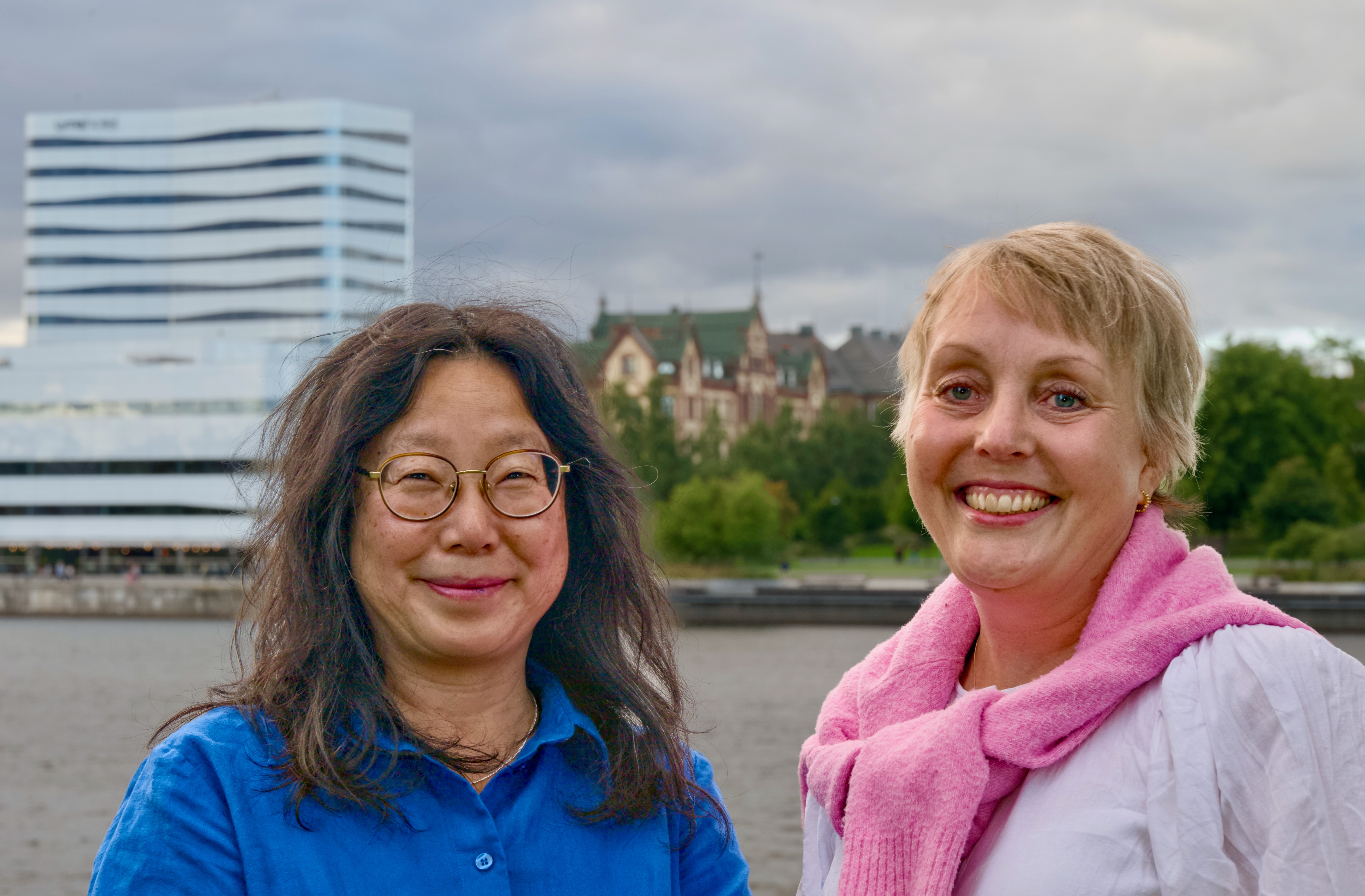 Maria Heimer och Nanna Forsgren ler utomhus nära vattnet, med moderna och historiska byggnader samt träd i bakgrunden under en molnig himmel.