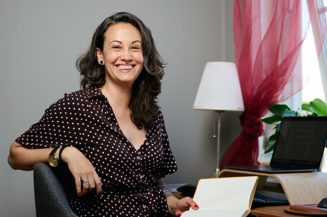 Nicole Galdíková i en brun prickig klänning sitter vid ett skrivbord, håller ett uppslaget anteckningsblock och ler; i bakgrunden syns en lampa, en växt och en bärbar dator.
