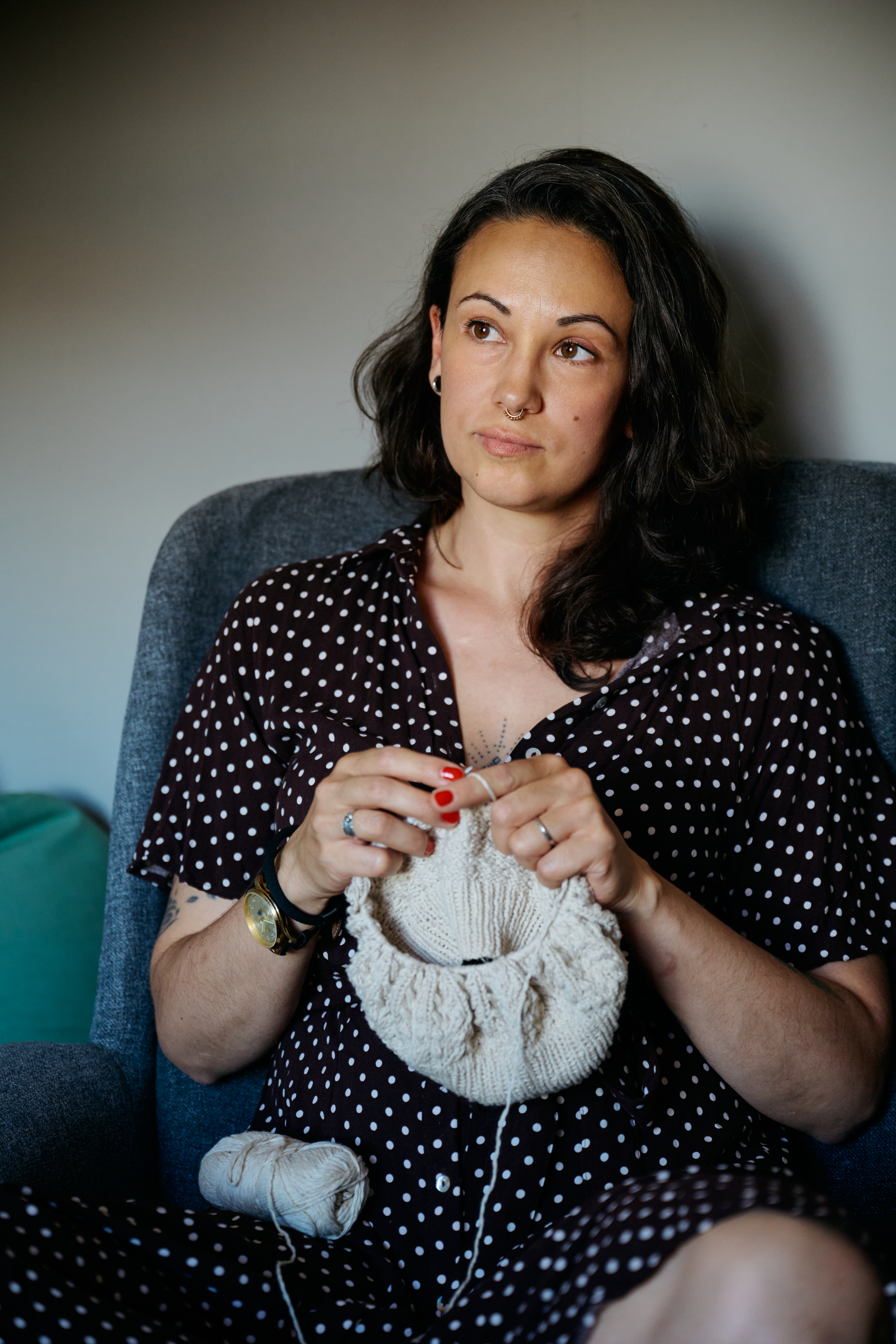 Nicole Galdíková i en prickig klänning sitter på en stol och håller i samt stickar med ett nystan ljusfärgat garn, vänd åt sidan.