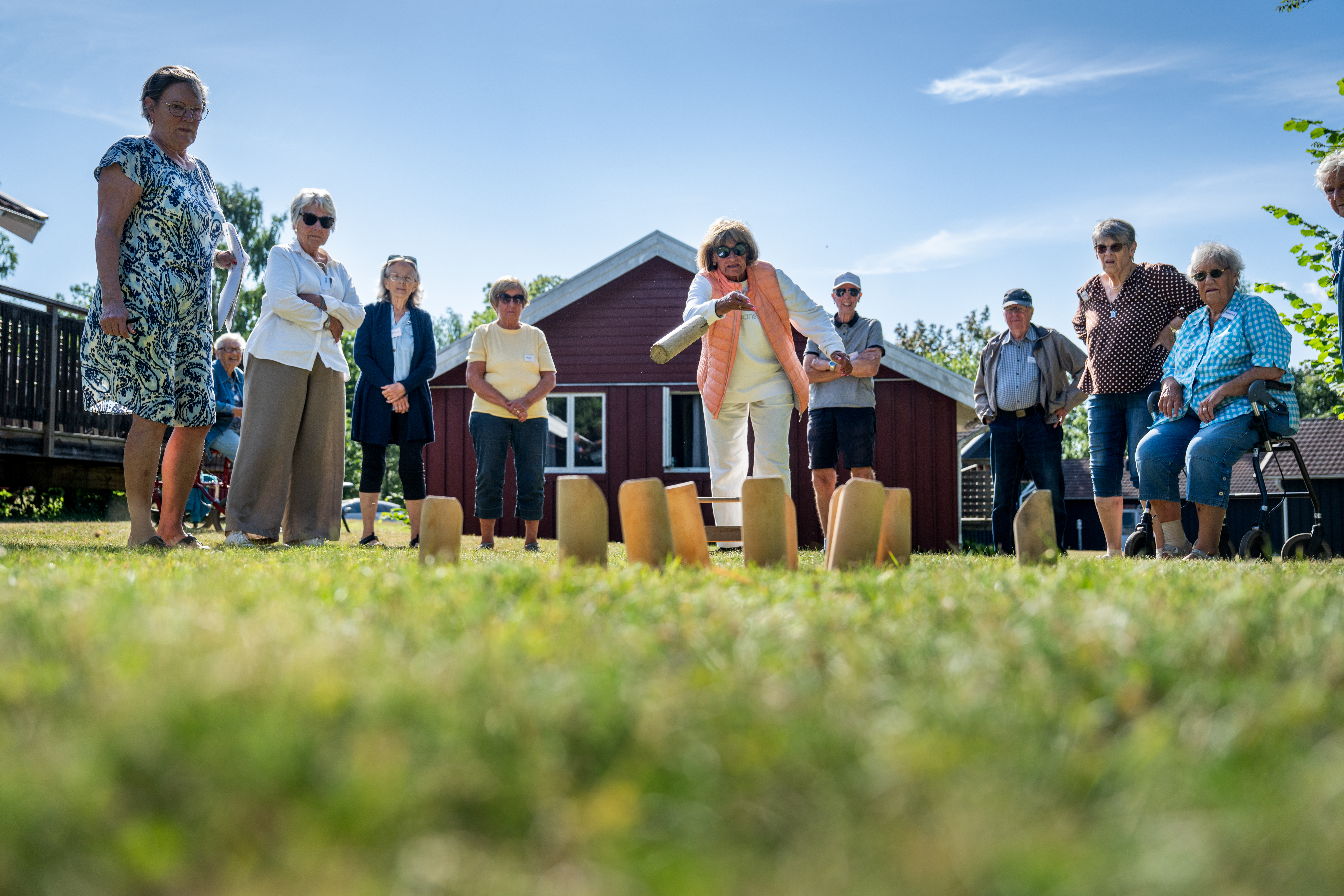 En grupp äldre vuxna står på gräset och tittar på en kvinna som kastar i en utomhusaktivitet, med ett rött hus i bakgrunden och himlen ovanför.