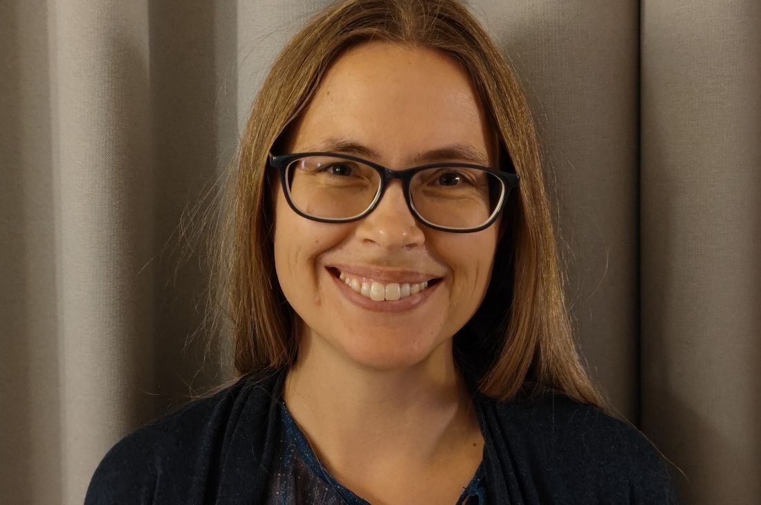 Malin Anderman med rakt brunt hår och glasögon ler mot kameran och står framför grå gardiner.