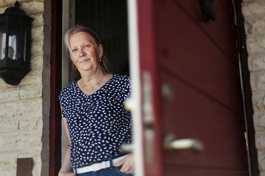 Kerstin Ingelmark med grått hår och en mönstrad blus står i en dörröppning med en delvis öppen röd dörr och en tegelvägg i bakgrunden.