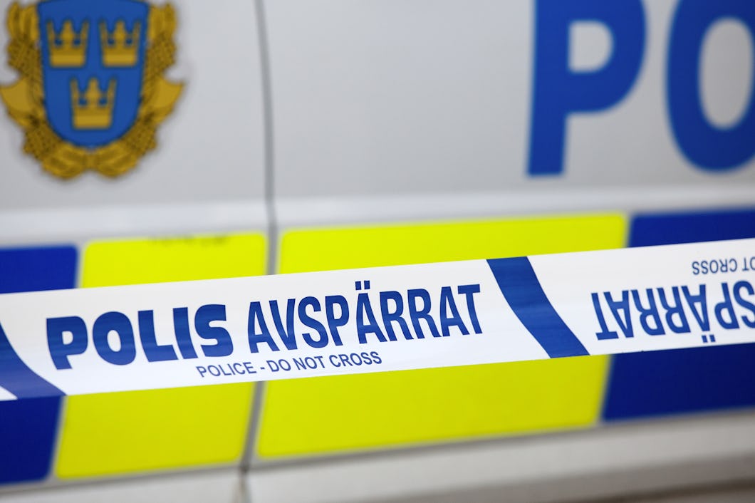 Närbild av svensk polisavspärrning med texten "POLIS AVSPÄRRAT - POLICE DO NOT CROSS" samt en del av ett polifordon i bakgrunden.
