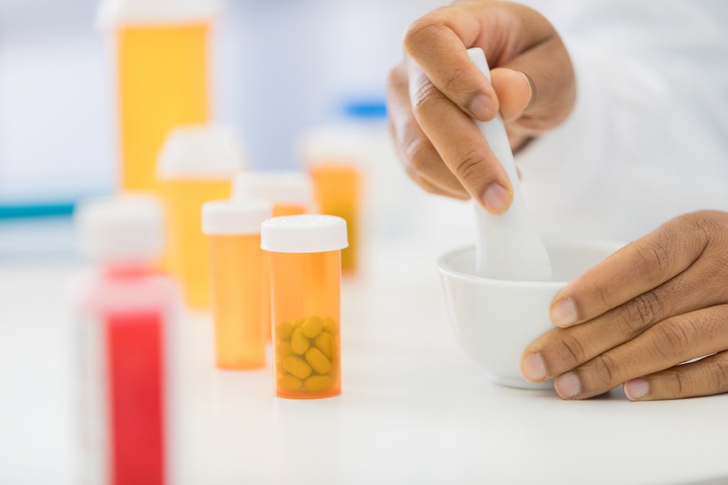 En person använder en mortel och mortelstöt för att mala något; flera medicinflaskor står på bordet.
