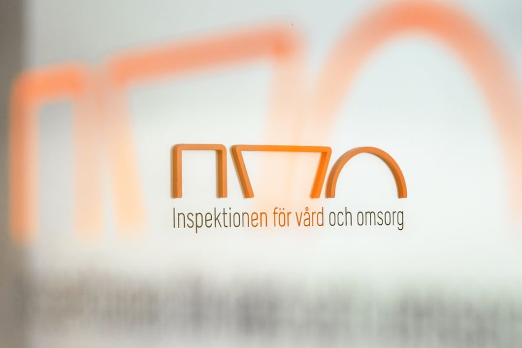 Logotyp och text på glas för "Inspektionen för vård och omsorg" i Sverige, med en abstrakt orange design ovanför texten.