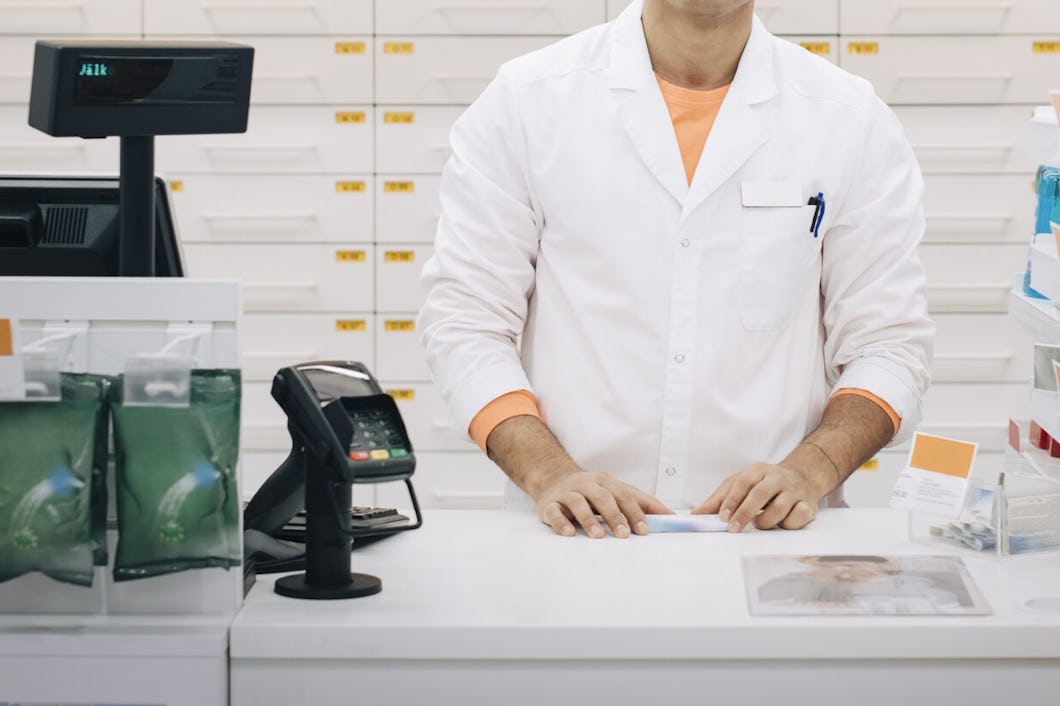 En farmaceut i vit rock står bakom apoteksdisken. I bakgrunden finns hyllor med mediciner.