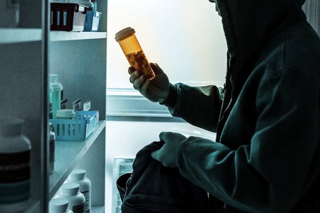 En person i en jacka med huva håller i en medicinflaska och lägger den i en väska i ett svagt belyst medicinskåp.