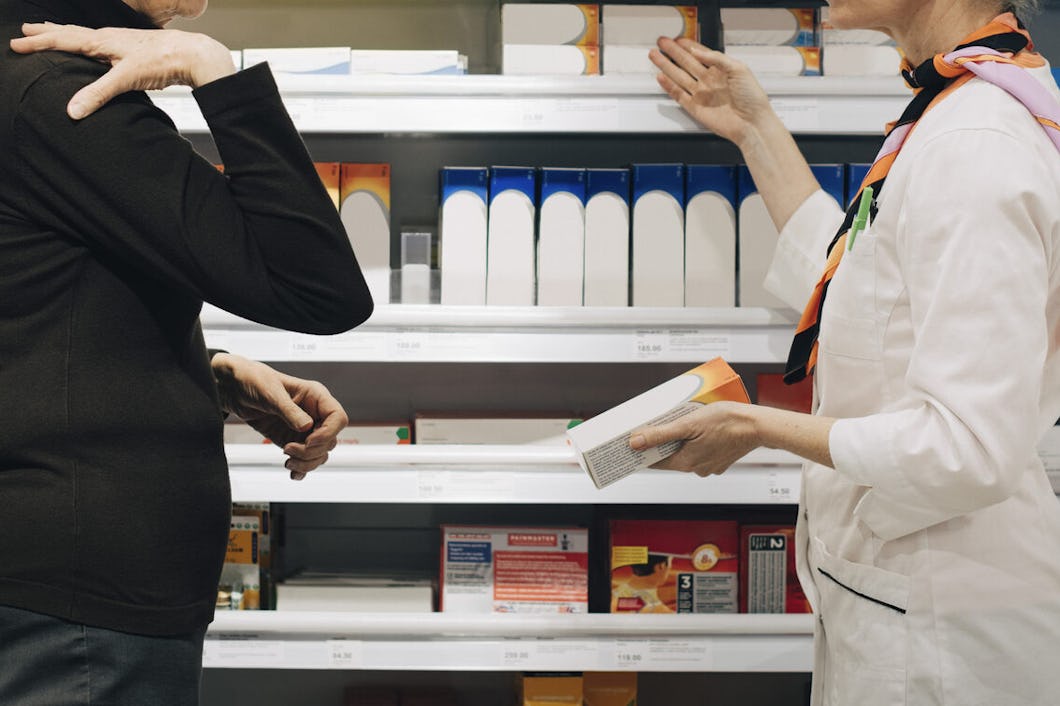 En person i ett apotek samtalar med en farmaceut som håller i en ask. I bakgrunden finns hyllor med flera produkter.
